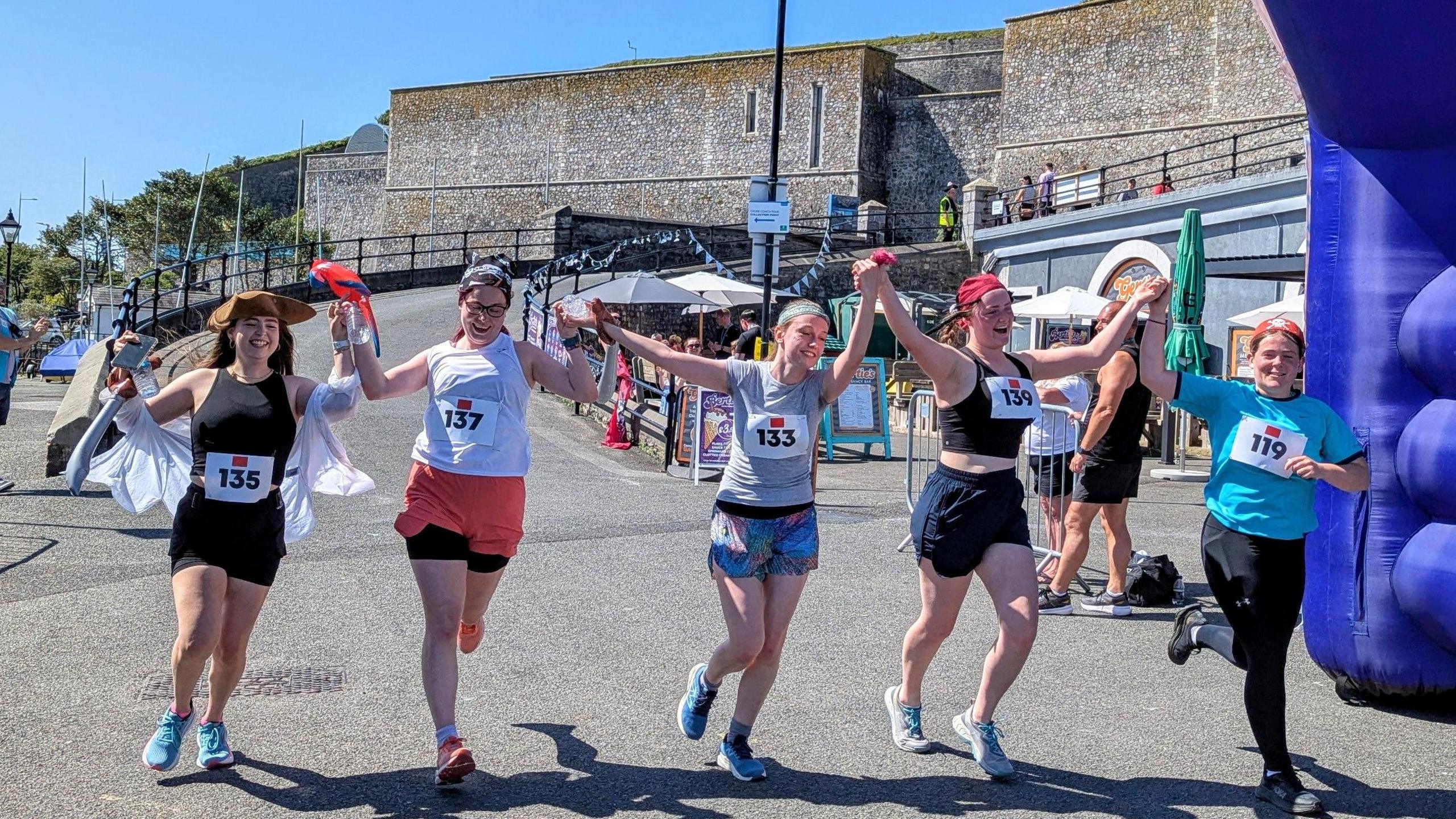 The finish line of a pirate fun run. There are five women running under a blue inflatable, they are wearing numbers pinned to their tops and wearing shorts, tank tops and trainers. The woman on the left is wearing a pirate hat and the other women are wearing bandanas.