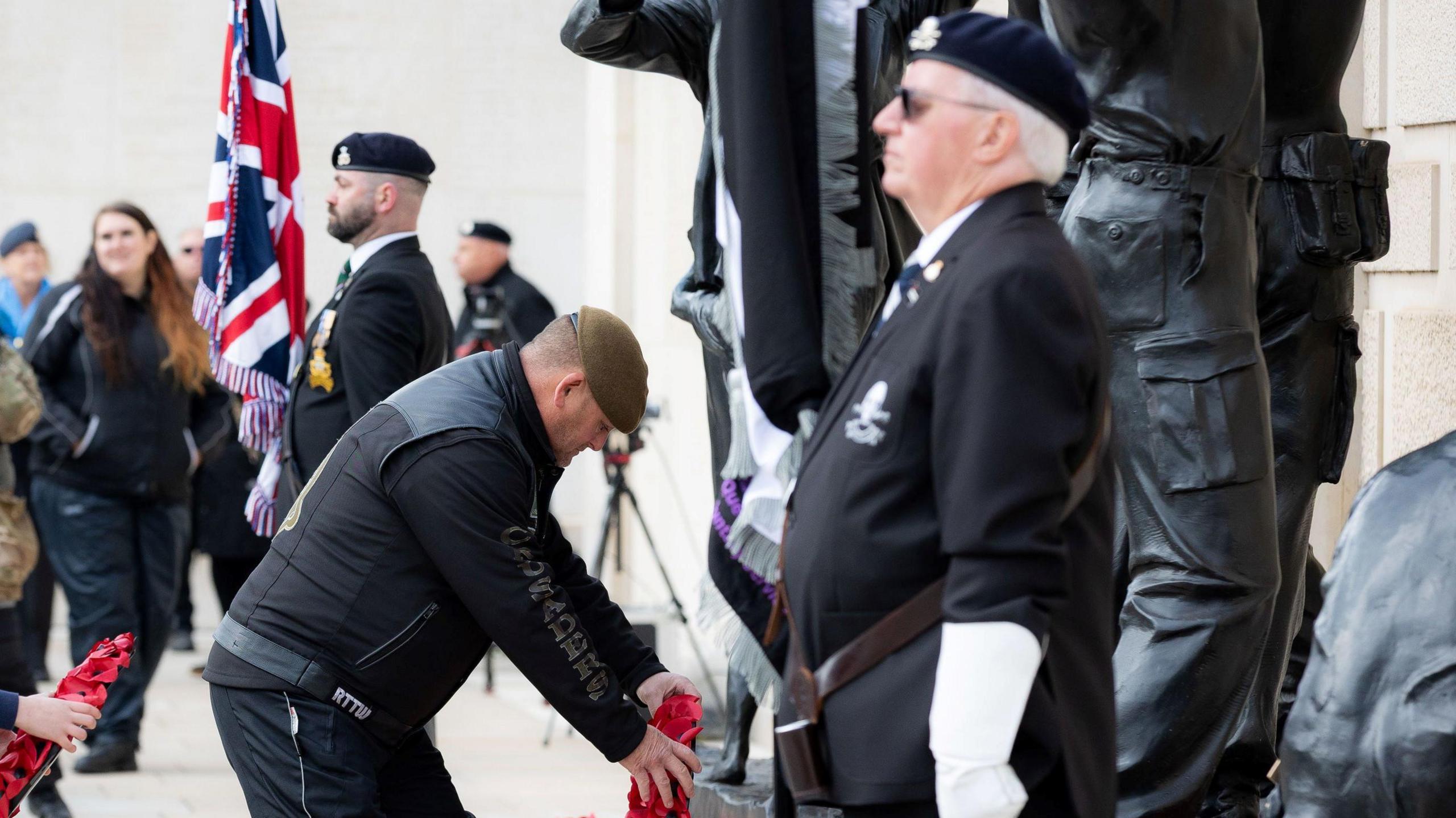 A man in a black jacket and black trousers is placing a wreath of poppies in front of him. In front of him are two military men standing straight and looking in front of them. A union jack which is rolled up.