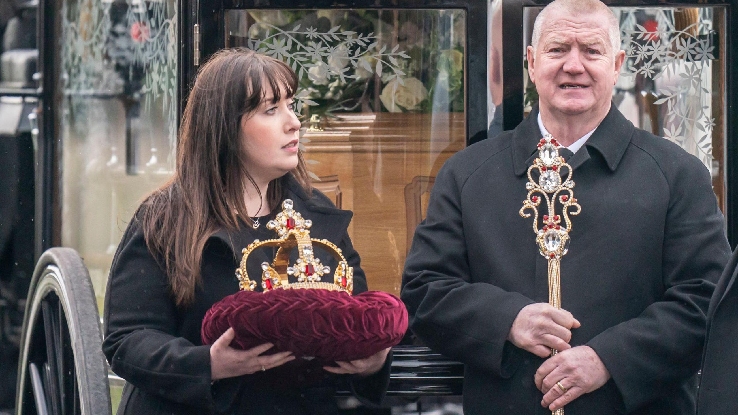 An ornately designed sceptre and matching golden crown - resting on a purple velvet cushion - which were won on RuPaul's Drag Race UK. They are being carried by a man and a woman into The Vivienne's funeral service.