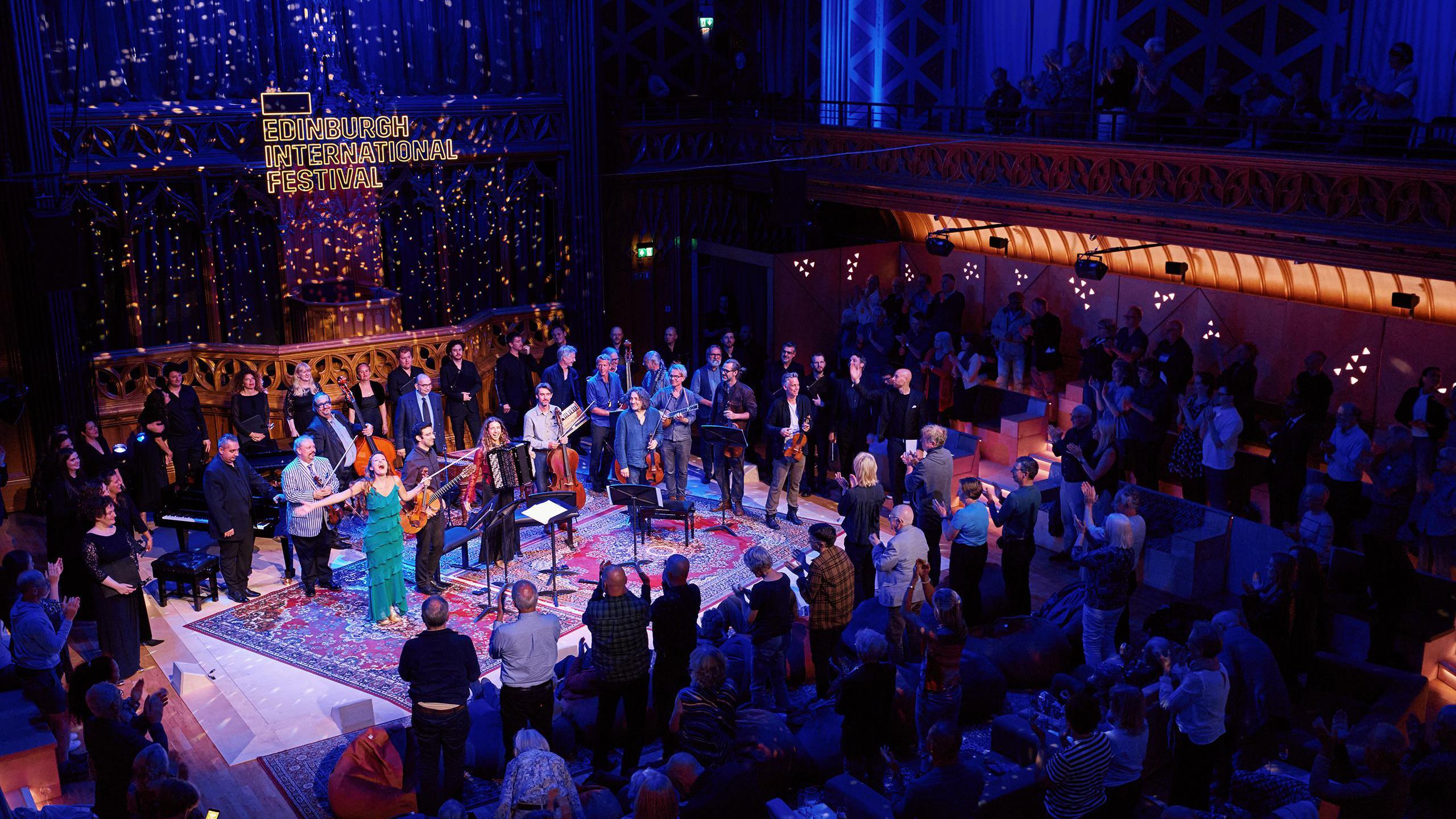 Wide shot of a live performance at the Edinburgh International Festival inside a grand, historic hall, with a large group of musicians and singers on a central stage surrounded by a standing audience, ornate balconies and patterned walls lit in blue and warm tones, and a glowing sign reading “Edinburgh International Festival” above the stage.