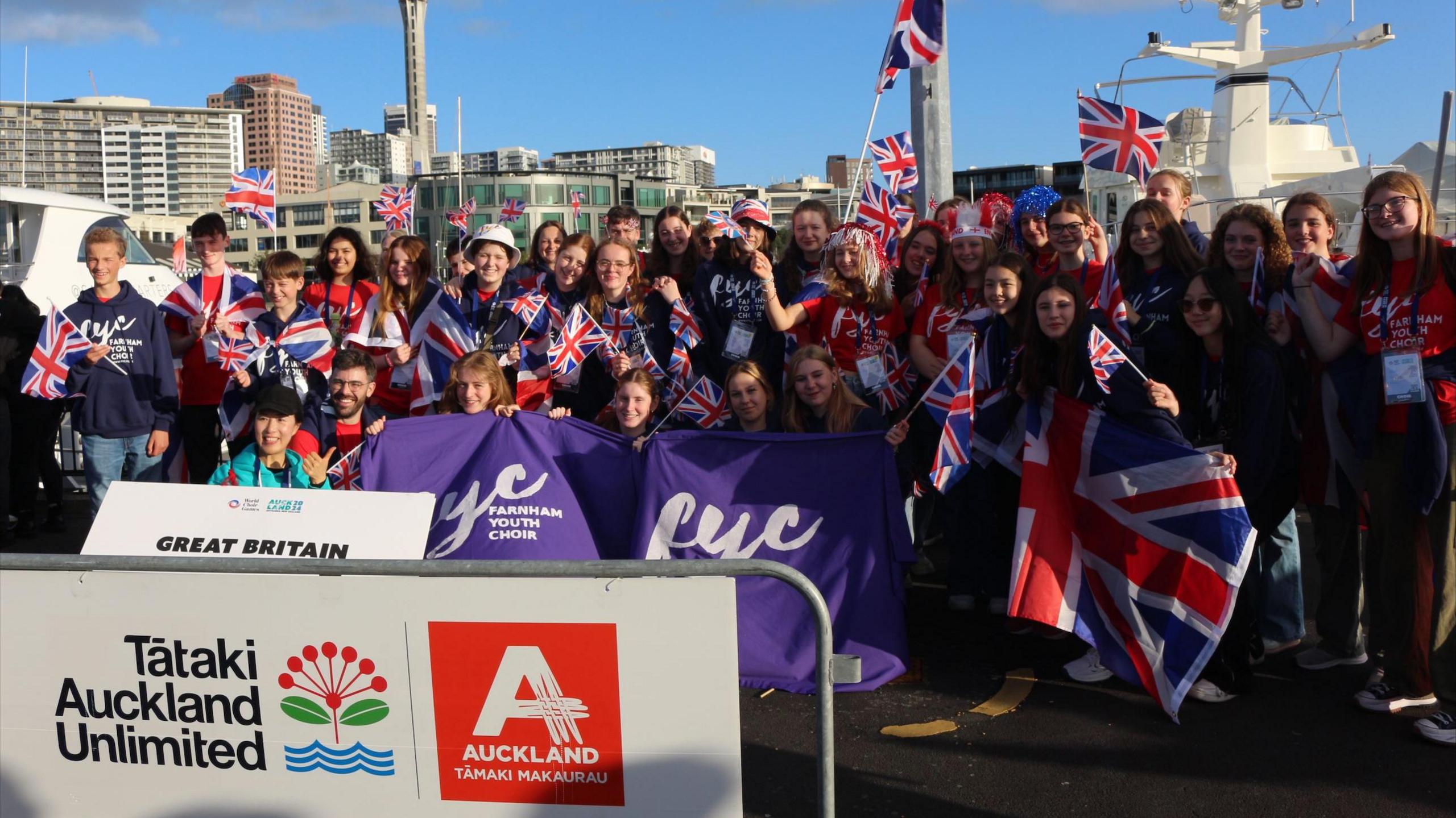 Members of the Farnham Youth Orchestra pose for a group photograph adorned in, and waving, Union Jack flags. They pose next to a white sign reading "Great Britain" and hold a purple banner with the 'FYC' logo on it.