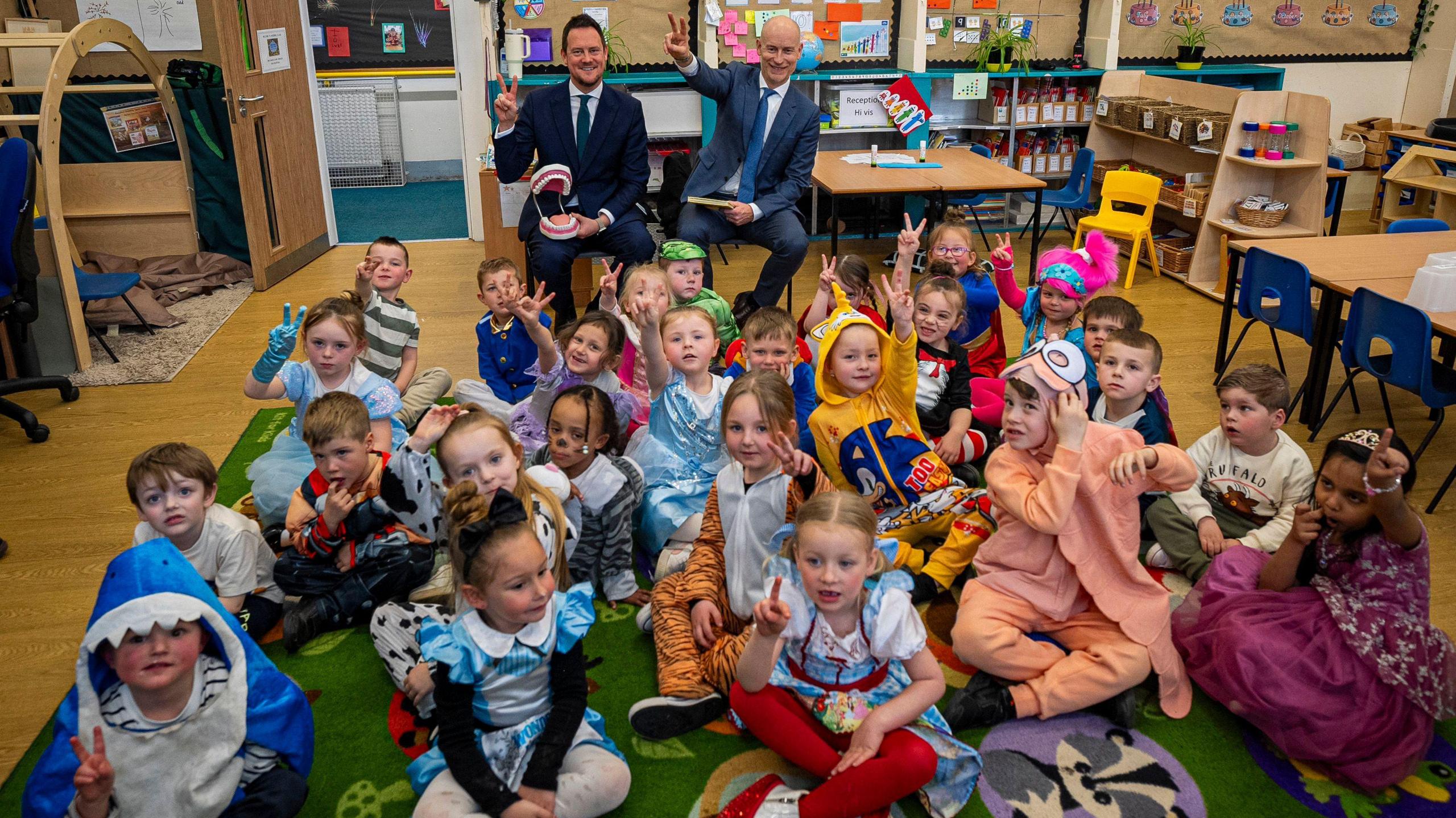 A large group of primary school pupils wearing World Book Day costumes at Fair Furlong Primary in Bristol wave at the camera. In the background on a seat are early education minister Stephen Morgan and care minister Stephen Kinnock