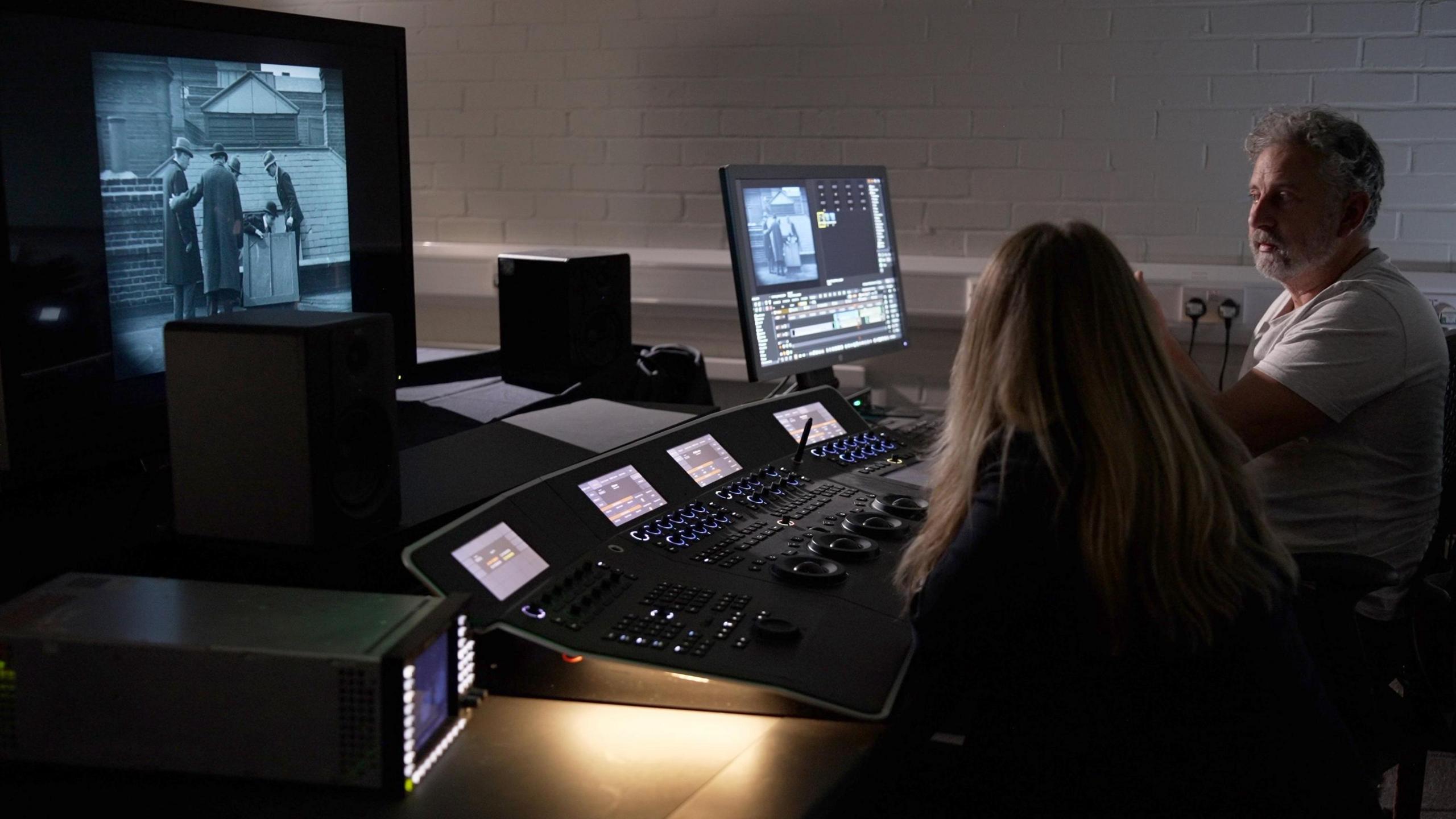 Two people sit in a dark room in front of a console with many lit buttons and a screen showing a Sherlock Holmes film