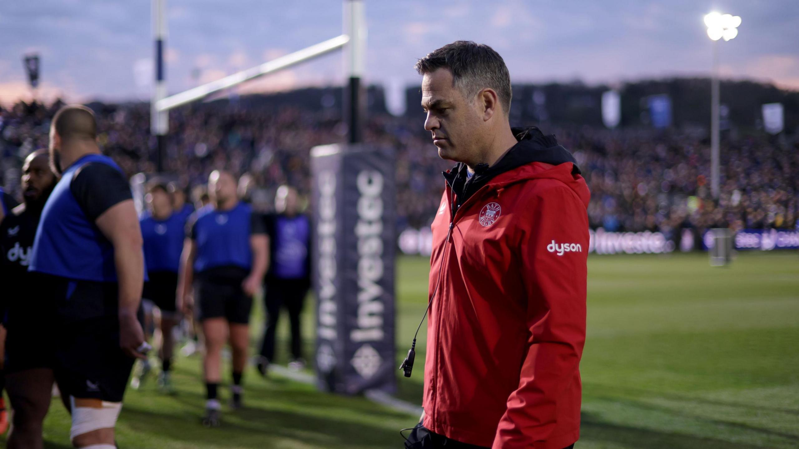 Johann van Graan walks on the pitch at the Recreation Ground with his head tilted down looking at the floor, as Bath players stand around the post in the background