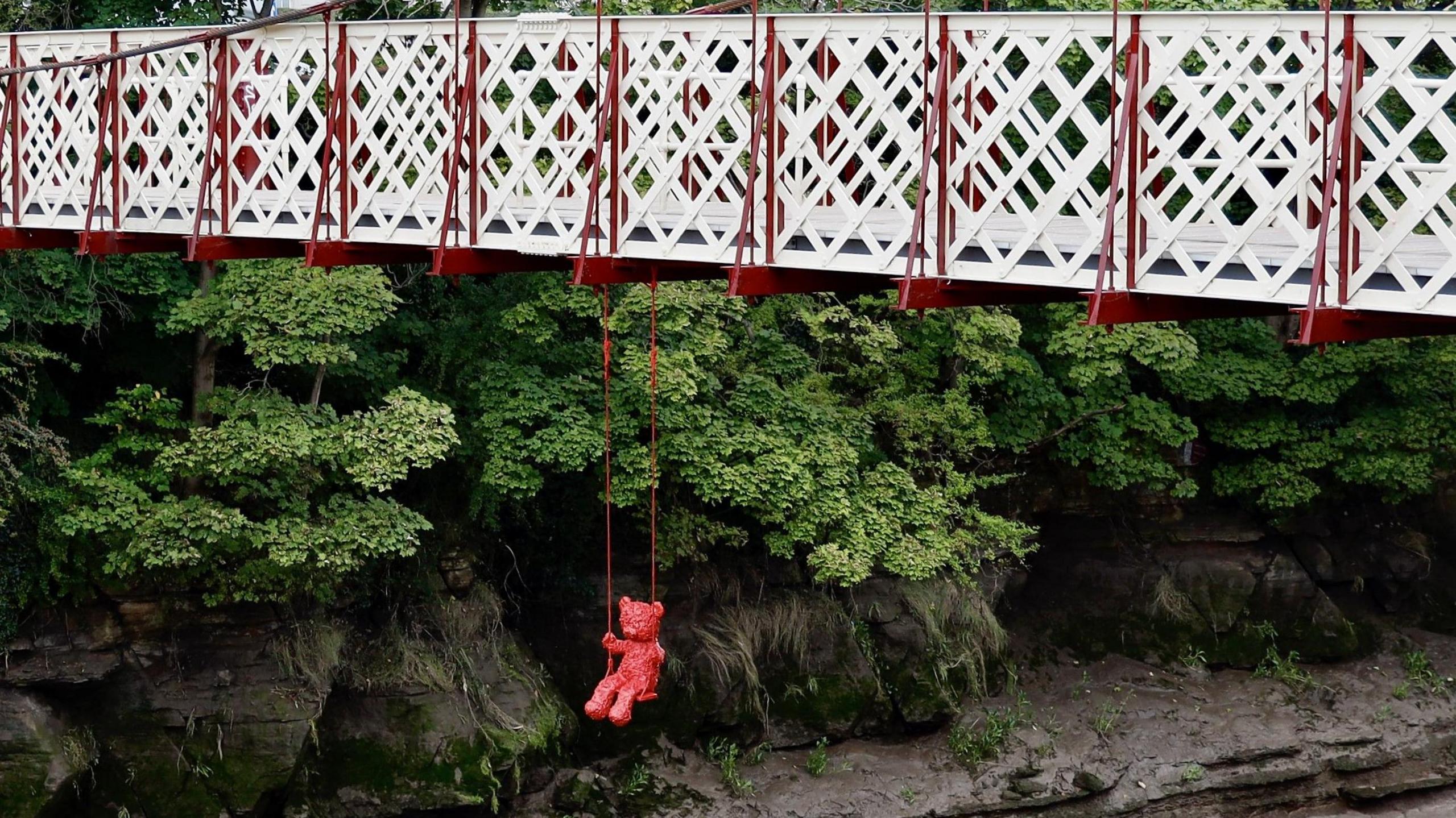 A red teddy bear street art creation hangs below Gaol Ferry Bridge in Bristol. The ropes holding the swing are also red, and in the background the muddy banks of the Avon and green trees and bushes on the banks are visible.