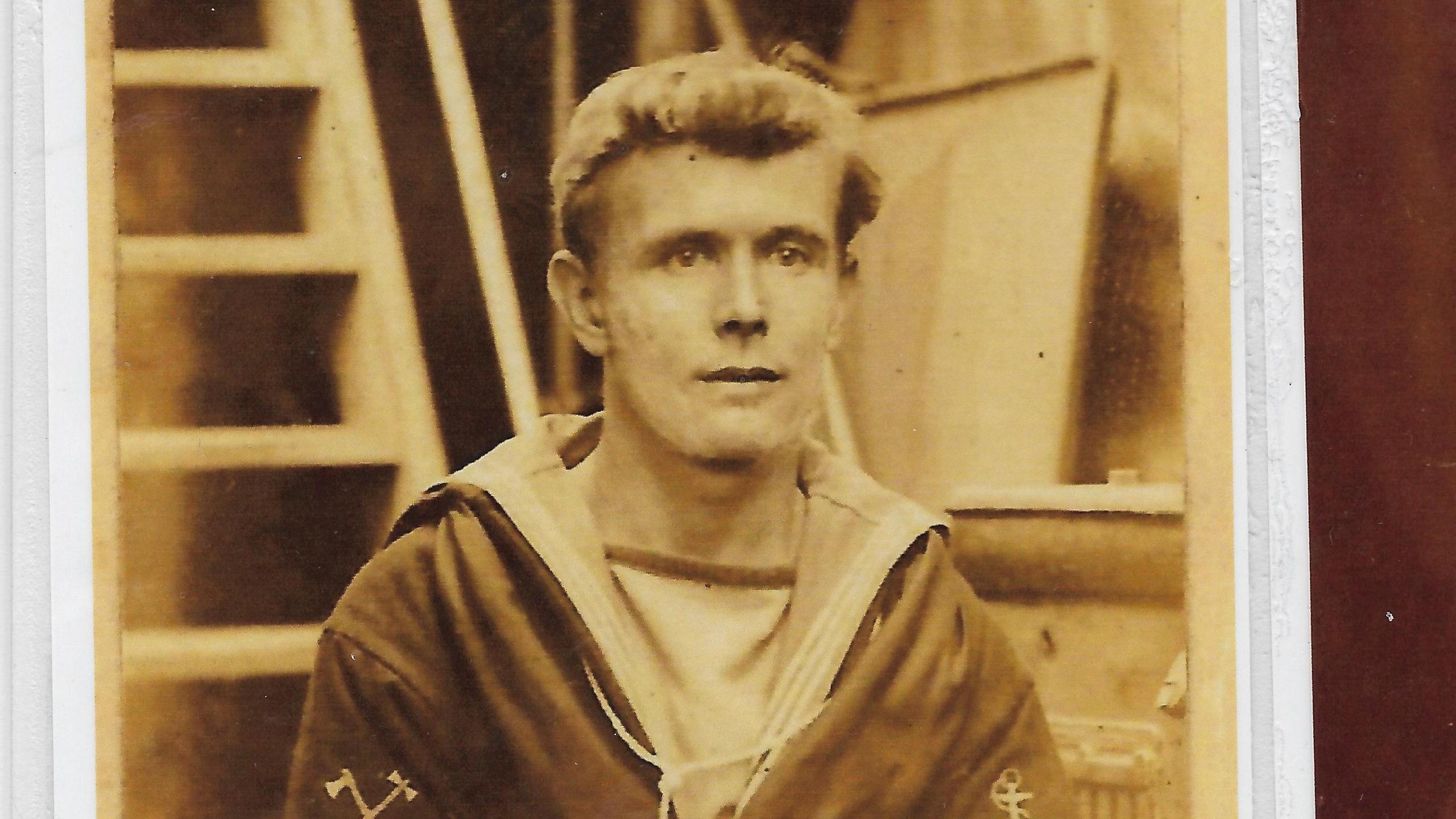 Black and white photo of Mr Barker wearing his Royal Navy uniform. He appears to be on a ship with stairs behind him. He appears young and has a slight quiff in his hair.