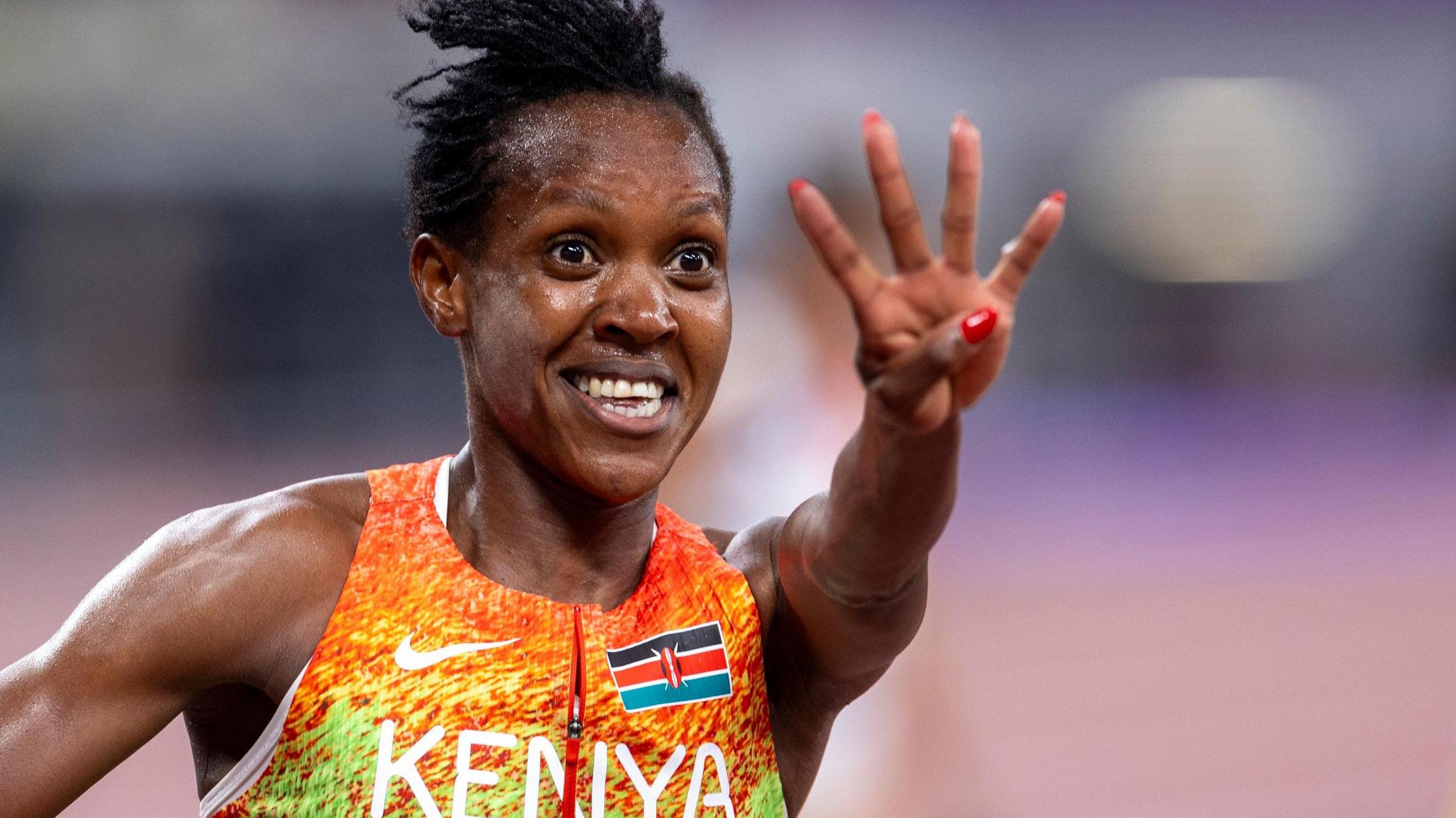 Faith Kipyegon, wearing a bright running vest with the word Kenya and Kenyan flag on it, smiles and extends her left arm to hold up four fingers as she celebrates winning 1500m gold at the World Athletics Championships in Tokyo