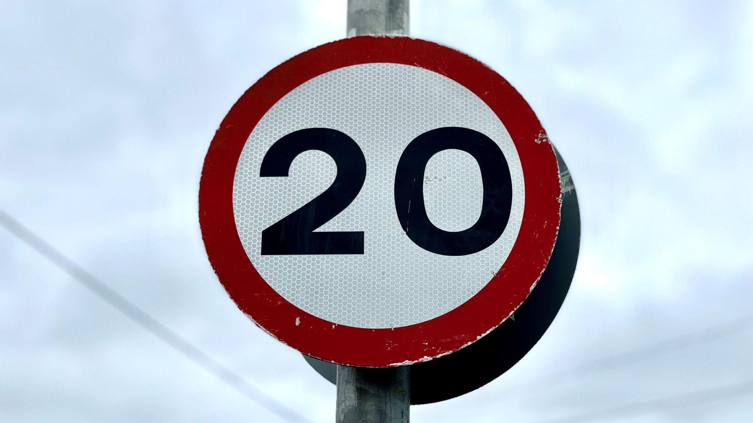 A close up of a circular 20mph sign - a black 20 on a white reflective background and a red rim on a steel pole