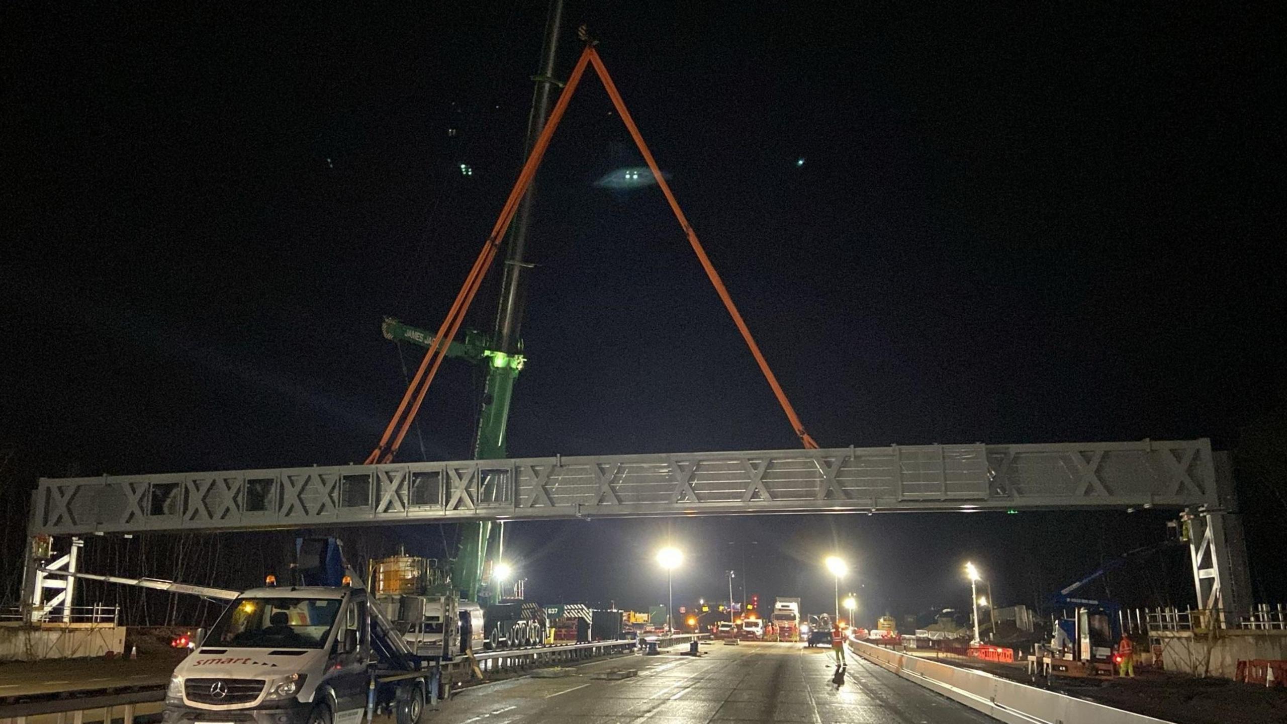 A night image of a large gantry being lowered in to place above the road with workers and vans on the road