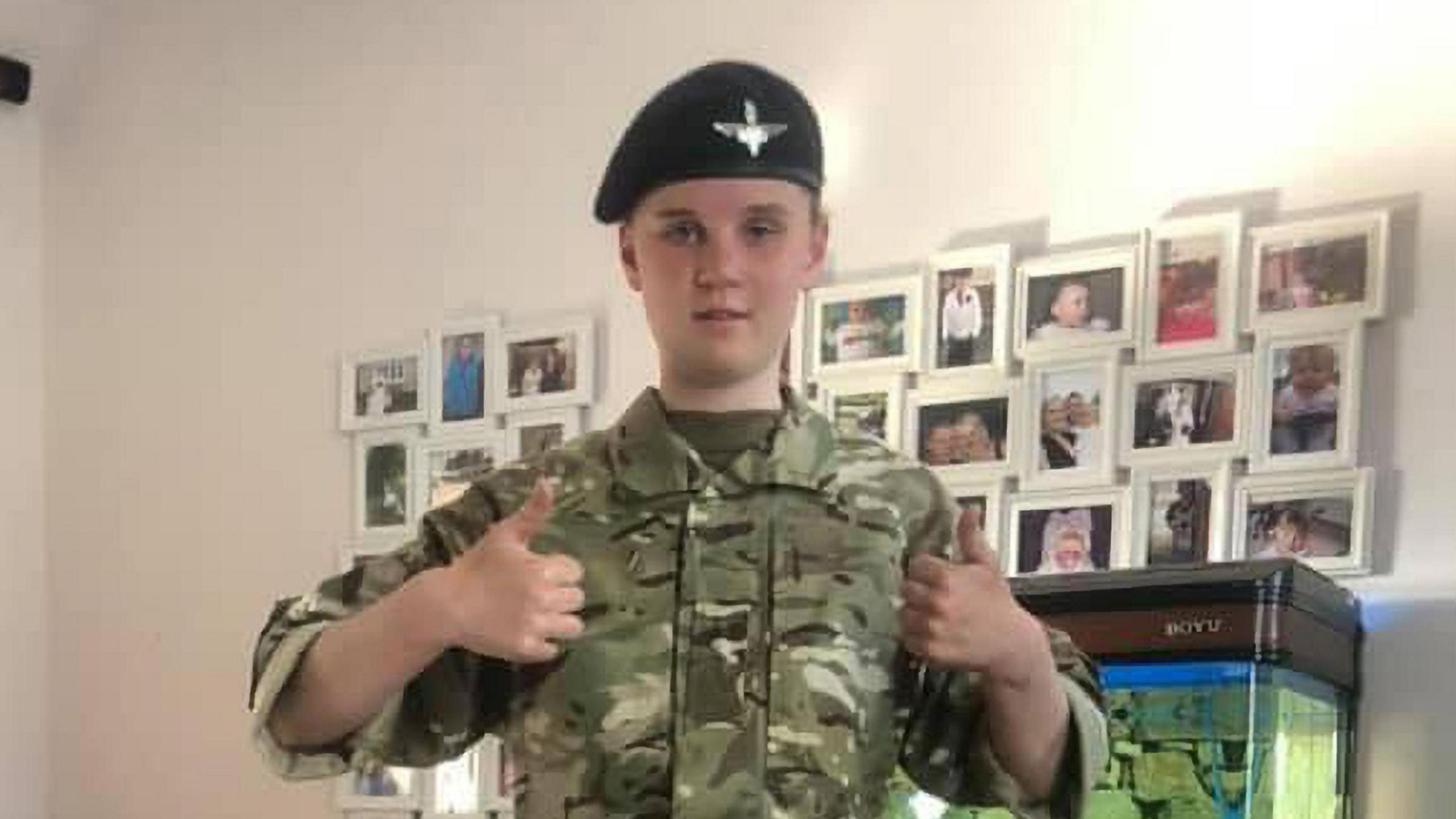 Holly Newton wearing army camouflage and a beret gives a double thumbs up to the camera. Behind her is a white wall with lots of pictures on it. 