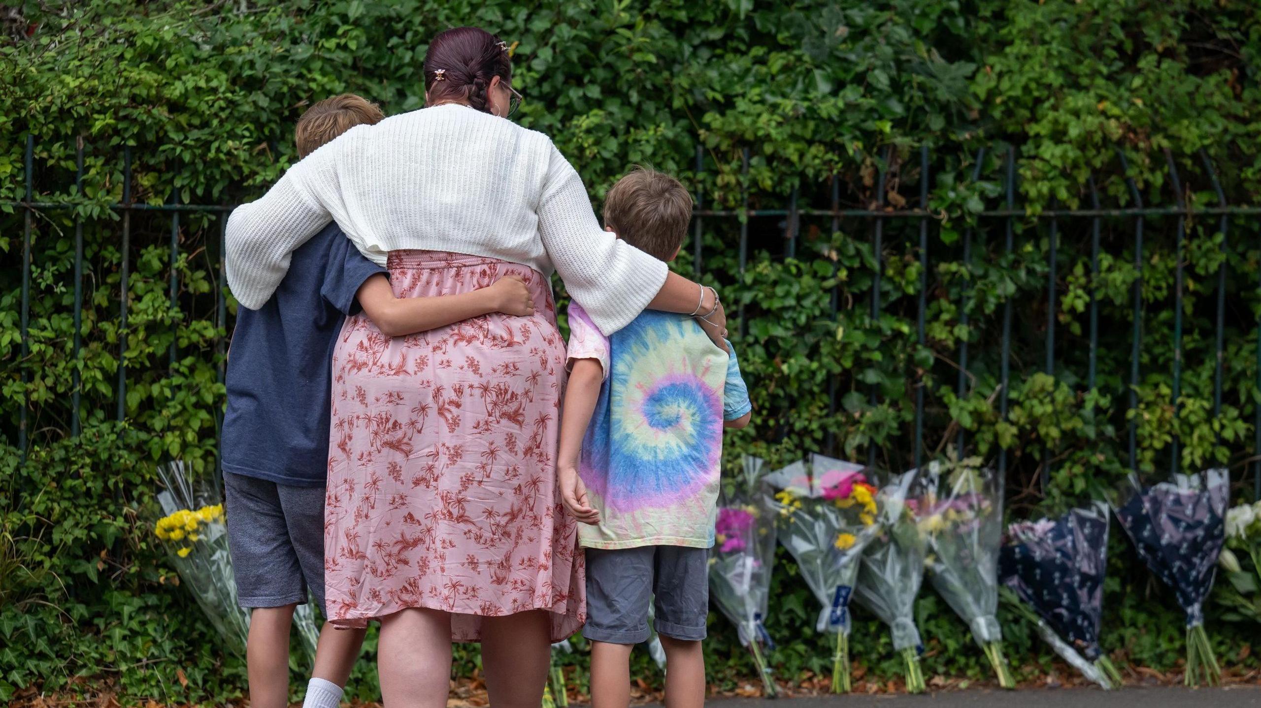 A woman, who wears a white cardigan and a pink floral dress, holds her arms around two boys. One on the left is taller, and wears a navy t-shirt and grey shorts. The boy on the right wears a tie-dye t-shirt and grey shirt. They are photographed from behind, as they look at a row of floral tributes against a hedge and a fence.