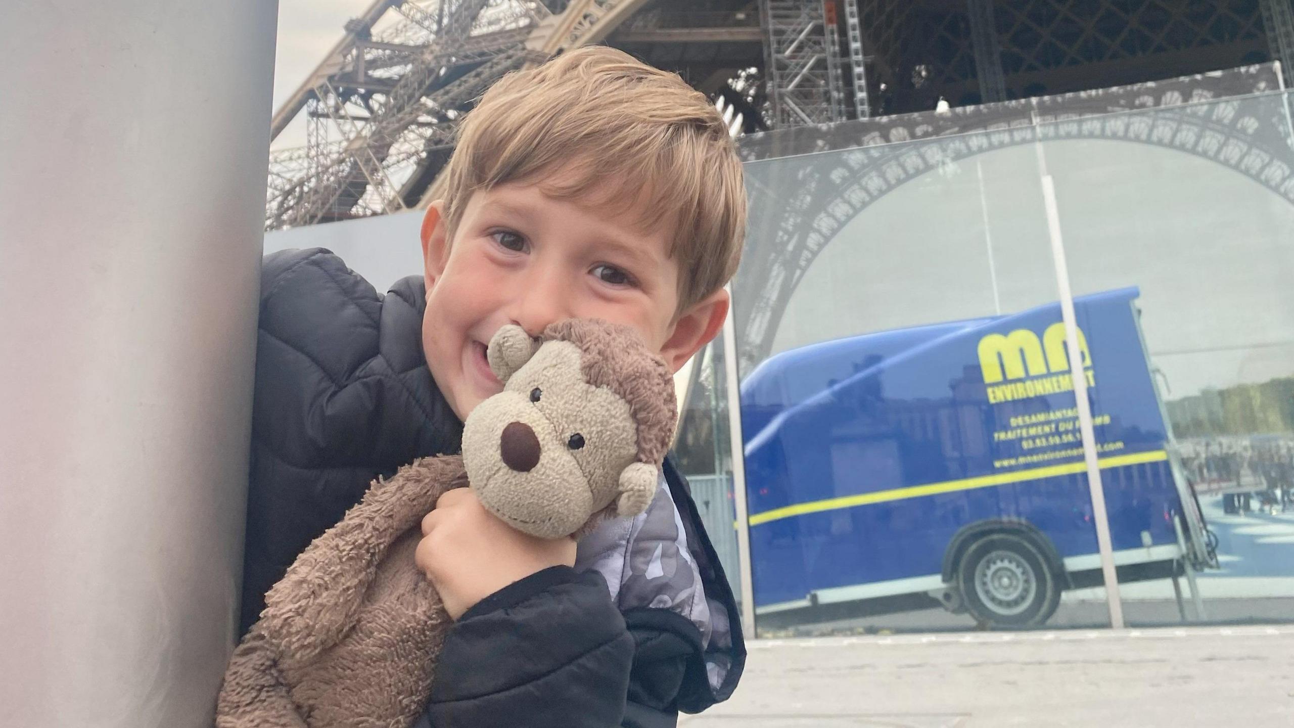 A young boy peeking round the corner of a grey post smiling in a cheeky way and holding a brown monkey teddy up to his cheek. He has dark blond hair and a fringe and is wearing a black coat. The bottom of the Eiffel Tower is in the background, as is a reflection of a blue van.