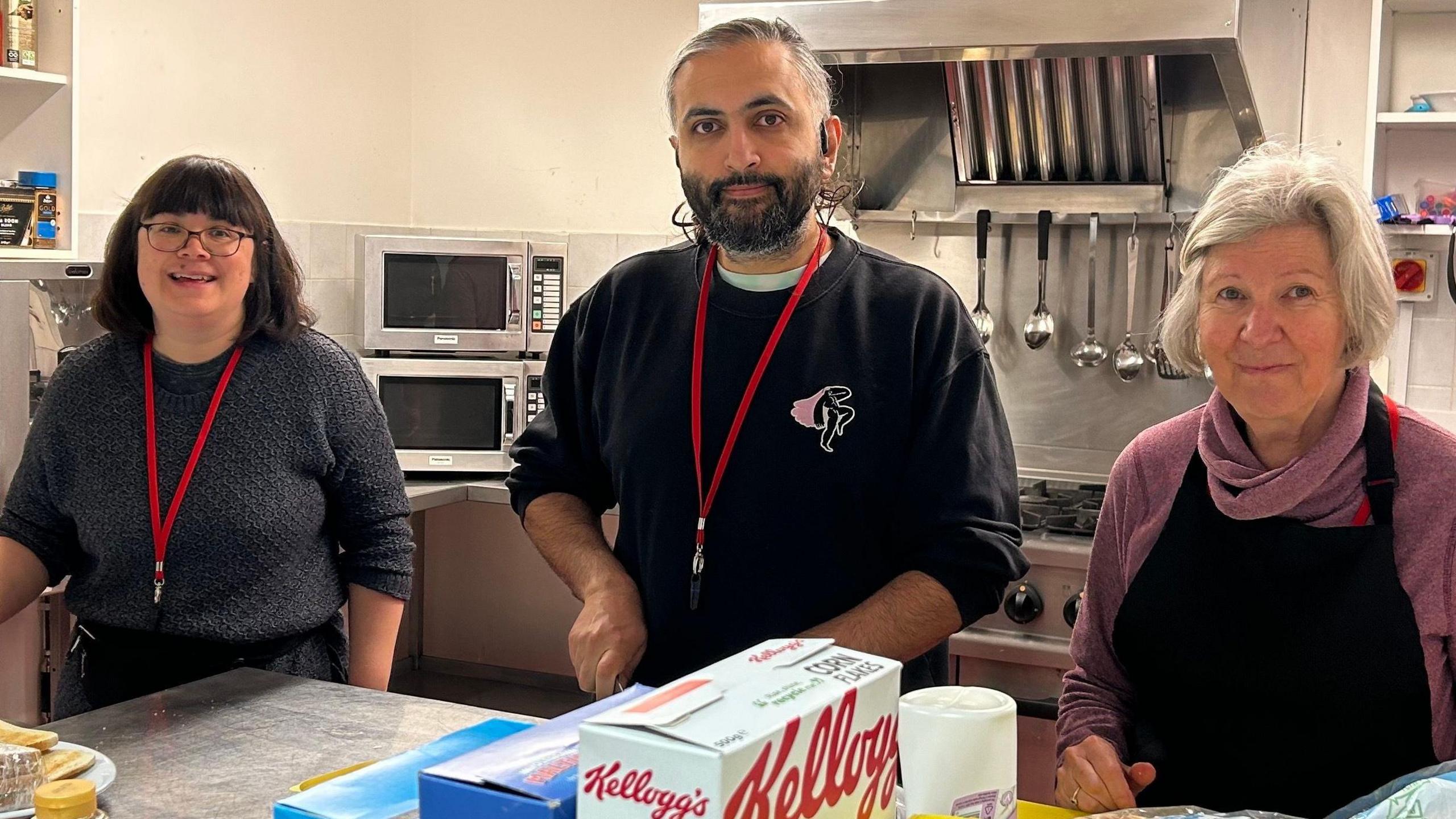 Three people, a woman, a man, and another woman, standing in a kitchen They all have red lanyards on. The woman on the right has dark shoulder length hair, with glasses and is wearing a dark jumper, the man in the middle has a dark jumper on and a woman on the right is wearing a black apron with a purple jumper underneath. They are in a kitchen, with a microwave, utensils and a cooker behind them. They are by a silver bench with food on it. 