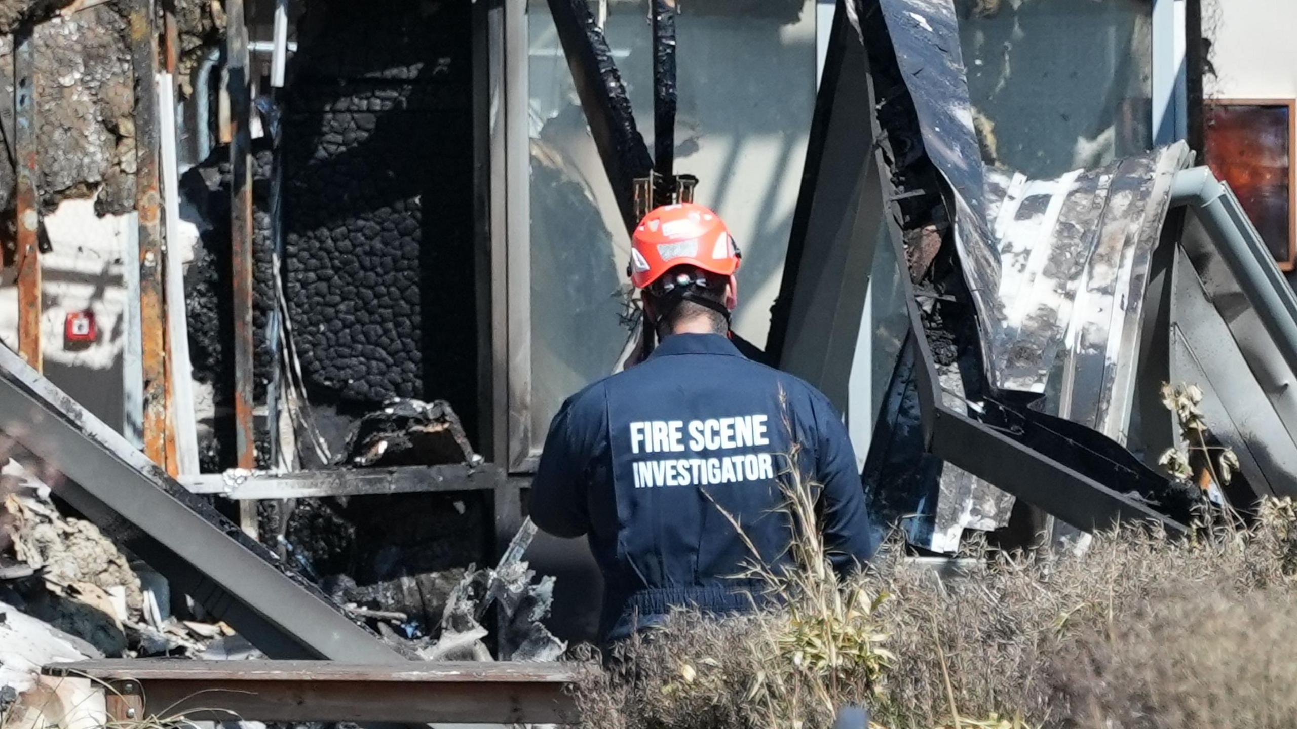 A fire scene investigator in a navy uniform and red helmet examines the charred remains of a severely burned building, surrounded by collapsed, blackened structure and scattered debris.