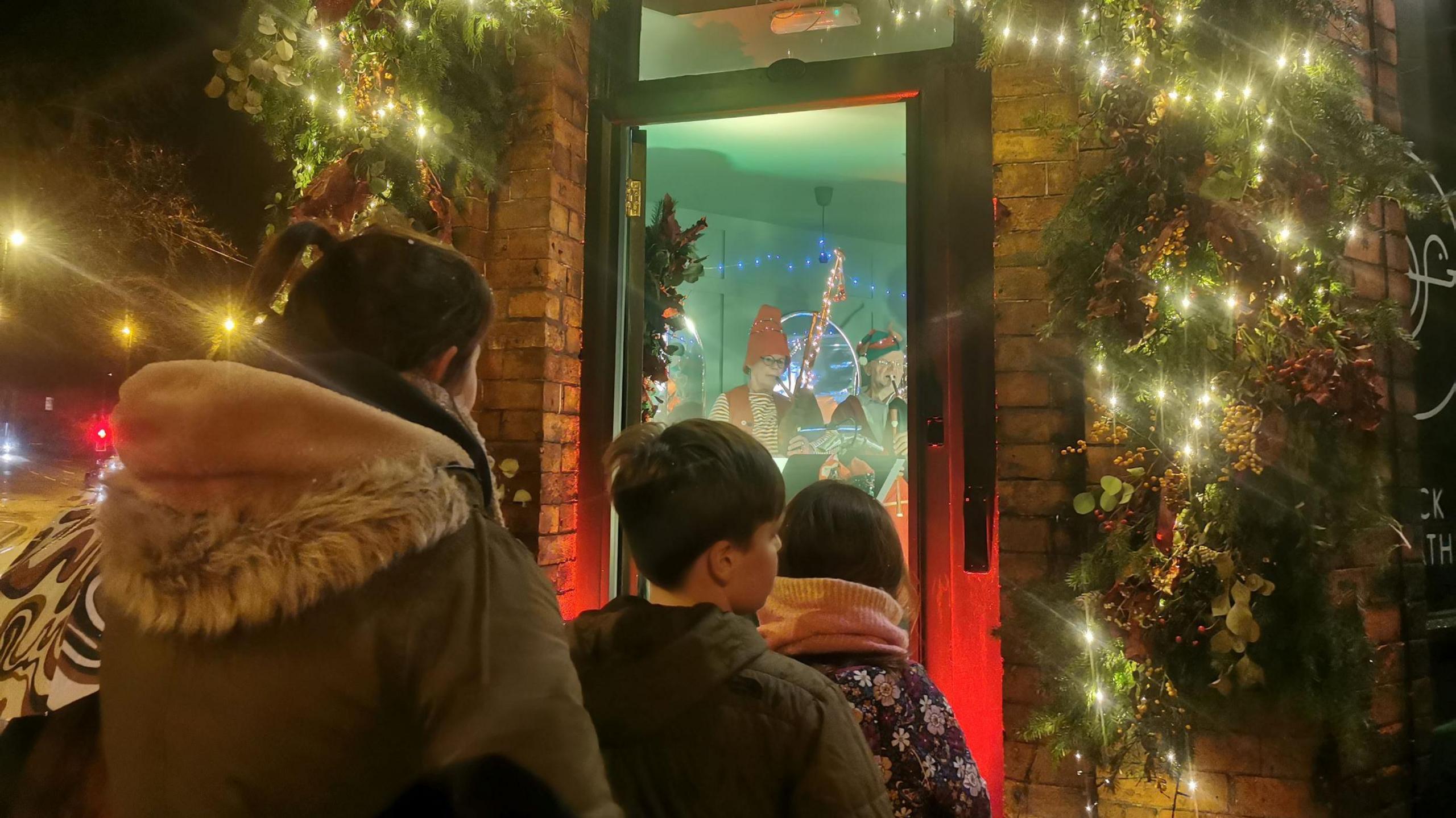 Three people stood in front of an open door with musicians stood inside the house which is covered in Christmas lights. Their backs are to the camera and they include a woman in a hooded coat with dark hair and two children in front of her with dark hair.