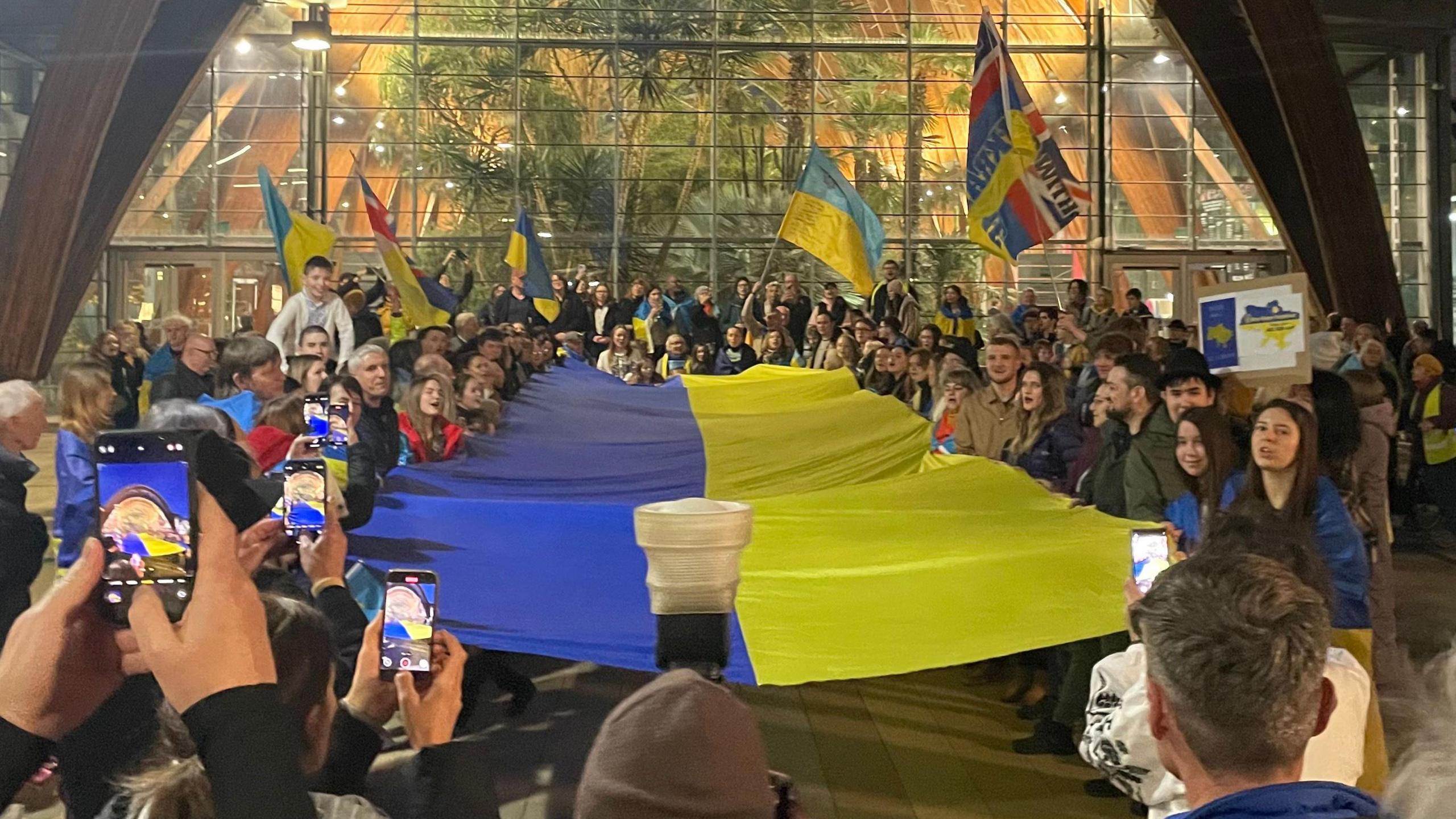 Attendees of the vigil hold a giant flag of Ukraine outside the Winter Garden. Some also have flags draped around their shoulders or are waving them in the air.