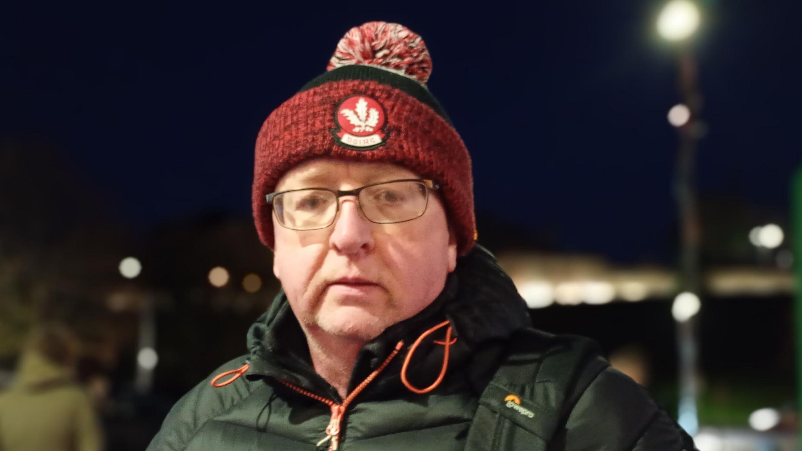 An older man wearing a red hat, bearing the insignia of Derry GAA, and a black coat and glasses. He has a neutral expression on his face and is standing on the street. In the background, out of focus, street lights can be seen.