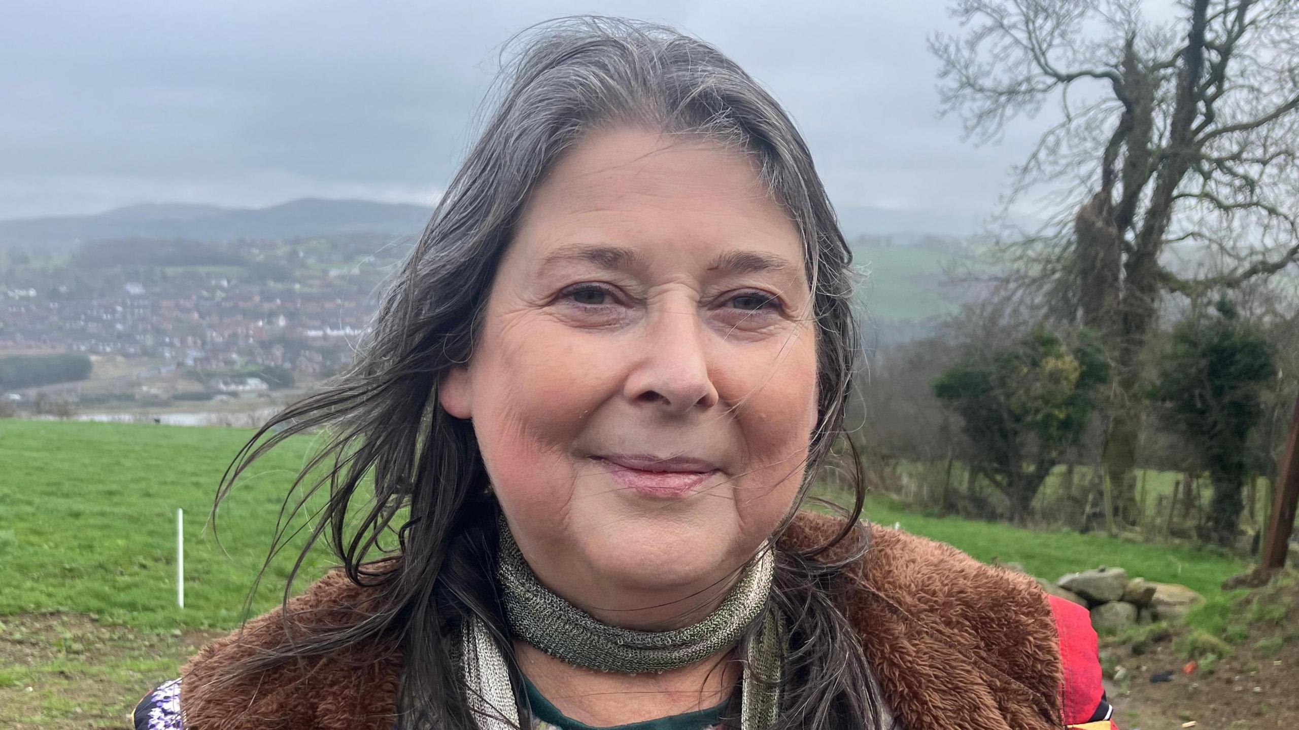 Ms Morgan with long black and grey hair, is wearing makeup and a brown fur jacket with a gold thin scarf.