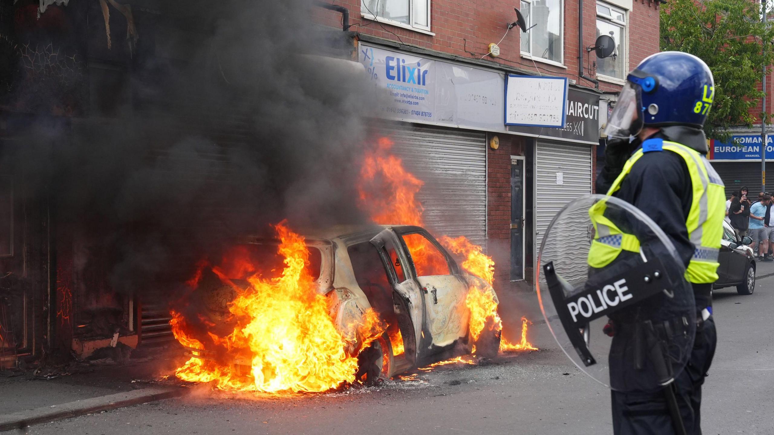 Car burning on a high street while a police officer looks on.