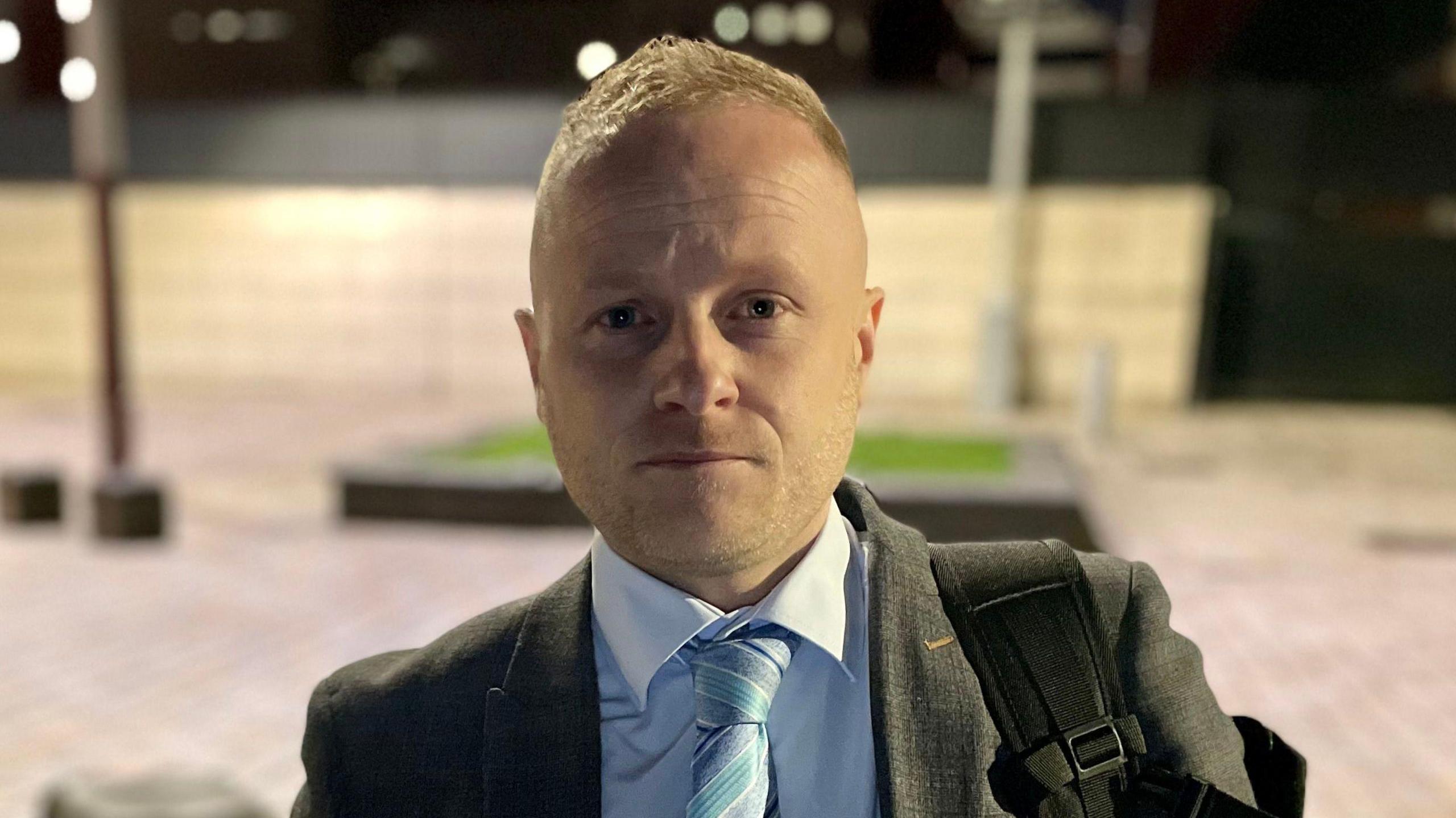 Jamie Bryson looking directly at the camera. He has short blonde hair, wearing a grey blazer, light blue shirt and blue striped tie, and carrying a backpack.