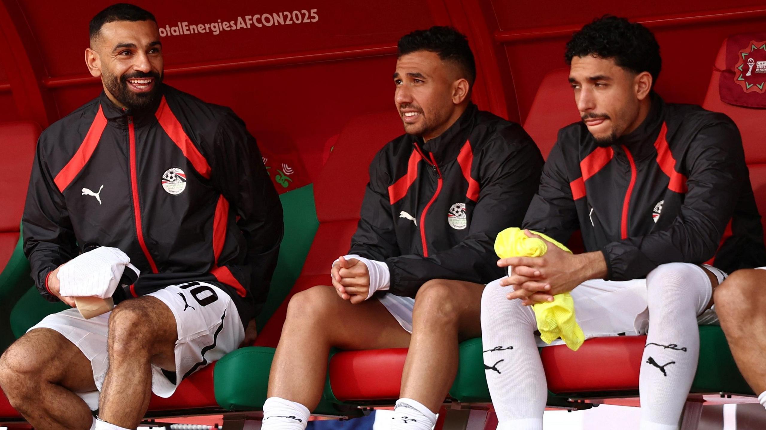 A photo of footballer Mohamed Salah smiling while sitting on the substitute bench with two team-mates for Egypt in the game against Angola at the Afcon 2025 football tournament. The players are sitting on red and green seats and wearing black tracksuit tops with red trim and white socks with black detailing.