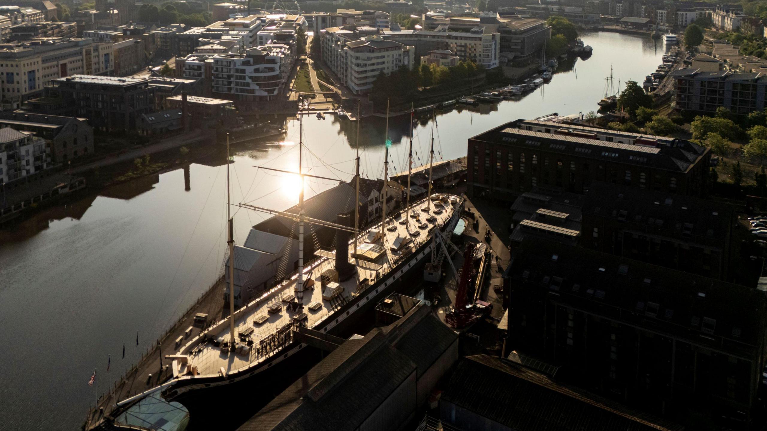 A drone view of Bristol's harbourside with the SS Great Britain in the foreground. The picture is taken on a sunny day and the sun can be seen reflected in the water