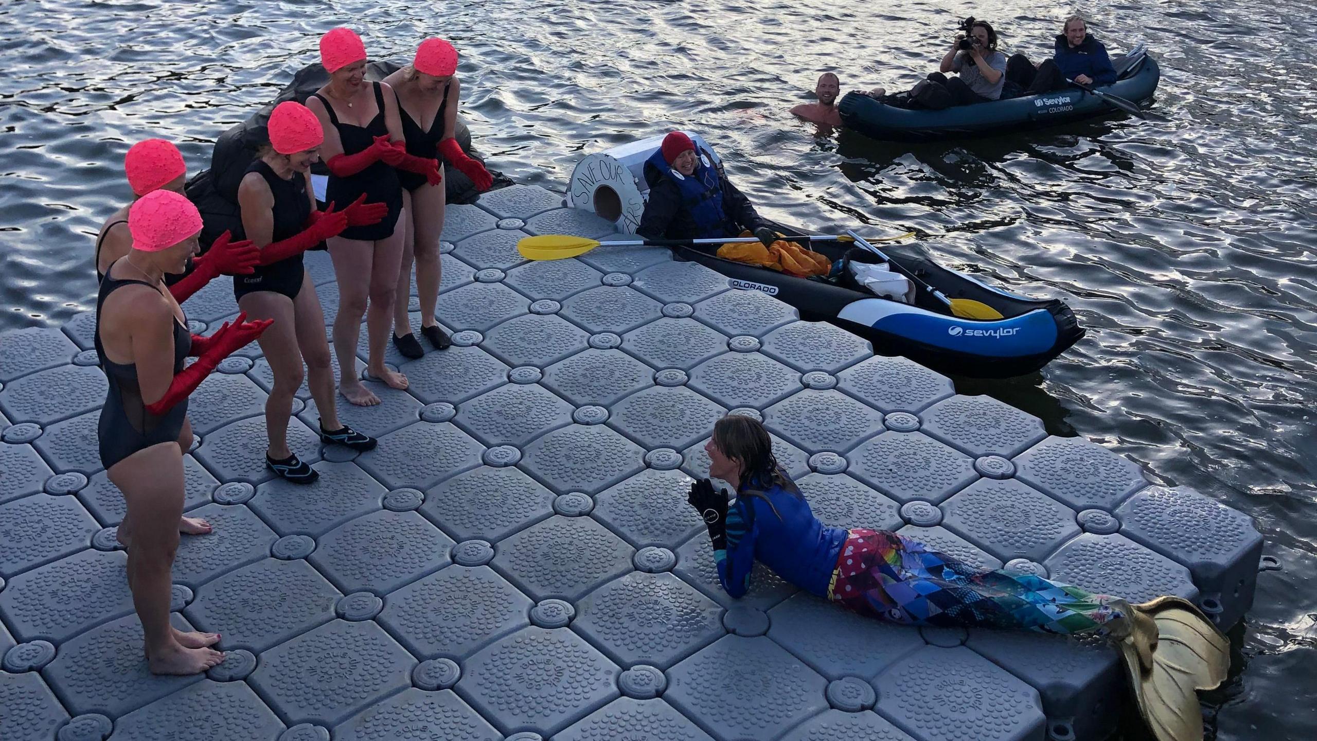 Woman in a mermaid costume lying on a platform in the water. She is surrounded by five women in black swimsuits, pink swimming caps and red gloves applauding her. In the water there are two paddle boats carrying people, one of which is taking a photo of the scene, and a person is in the water holding on to the paddle boat on the right. 