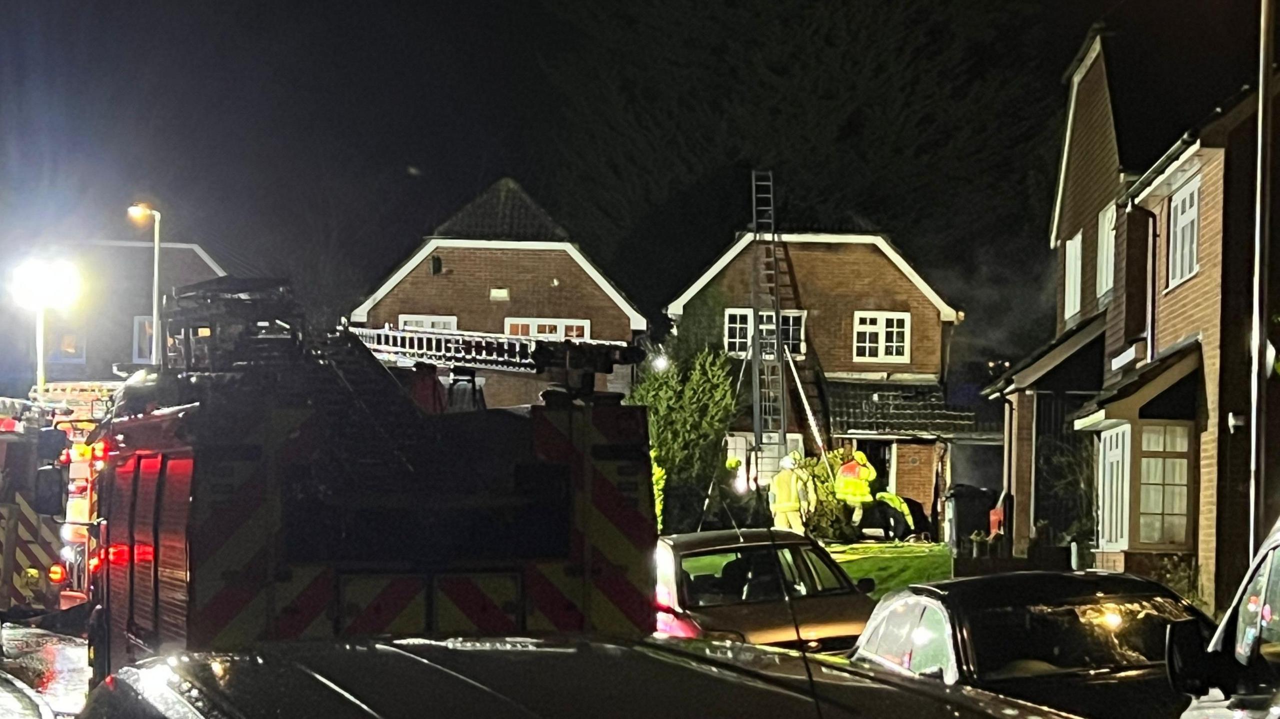 A fire engine and the back of a second engine seen attending a house fire in Frenches Farm Drive, Heathfield. Personnel can be seen at the house in high-viz gear, while a ladder is propped against the front of the house and extends onto the roof.