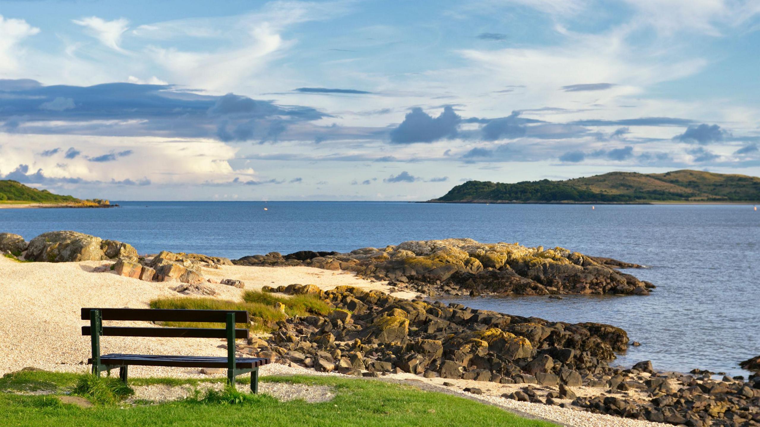 A bench looking out to sea across the Solway Firth with rocks jutting out into the water and green hills on the horizon