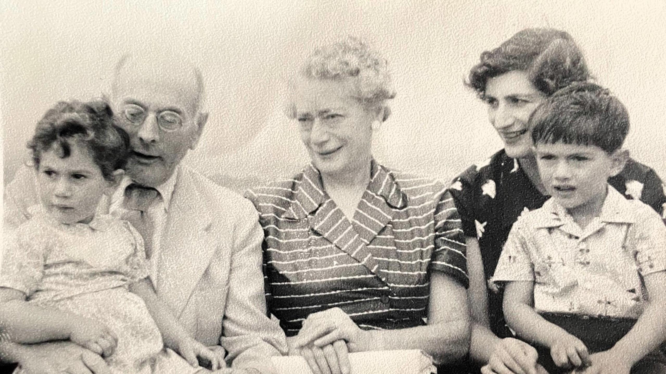 Llun du‑a‑gwyn o deulu Syr Michael Moritz yn eistedd mewn rhes, gyda phum person o wahanol oedrannau. Mae'r dillad â phatrymau nodweddiadol o’r 1950au, gan gynnwys siwt ysgafn, ffrog streipiog a chrysiau printiedig.