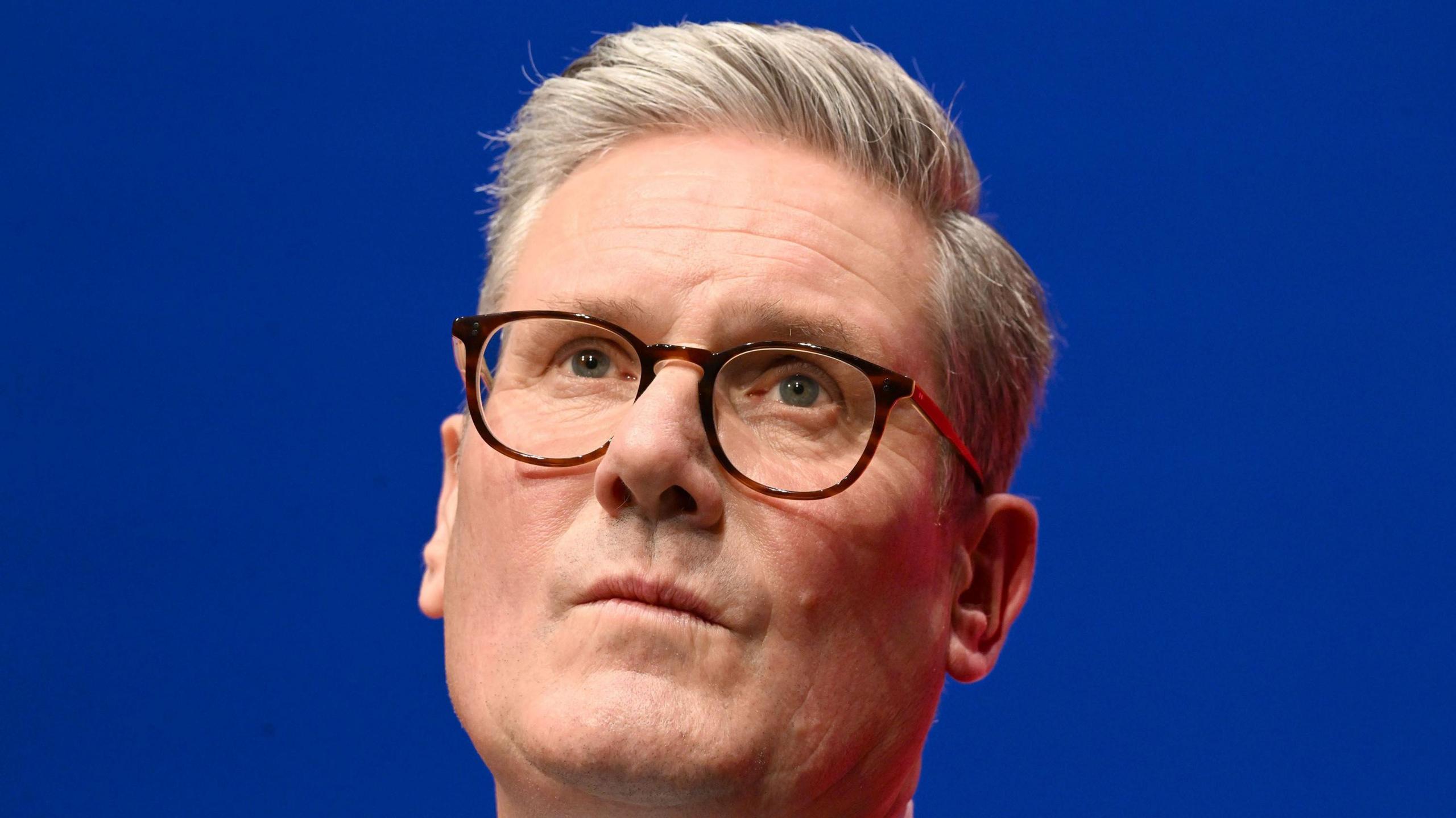 Sir Keir Starmer wearing black rimmed glasses at a conference in Liverpool, with a blue background 