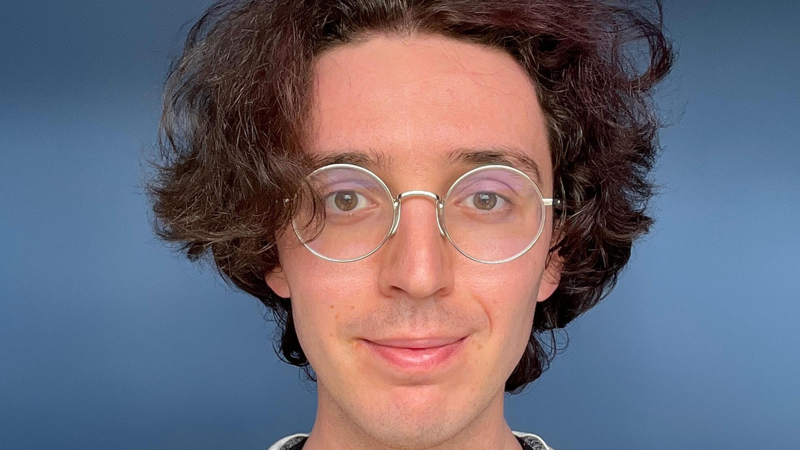 A picture of a person's face with brown wavy hair and circular glasses in front of a blue background