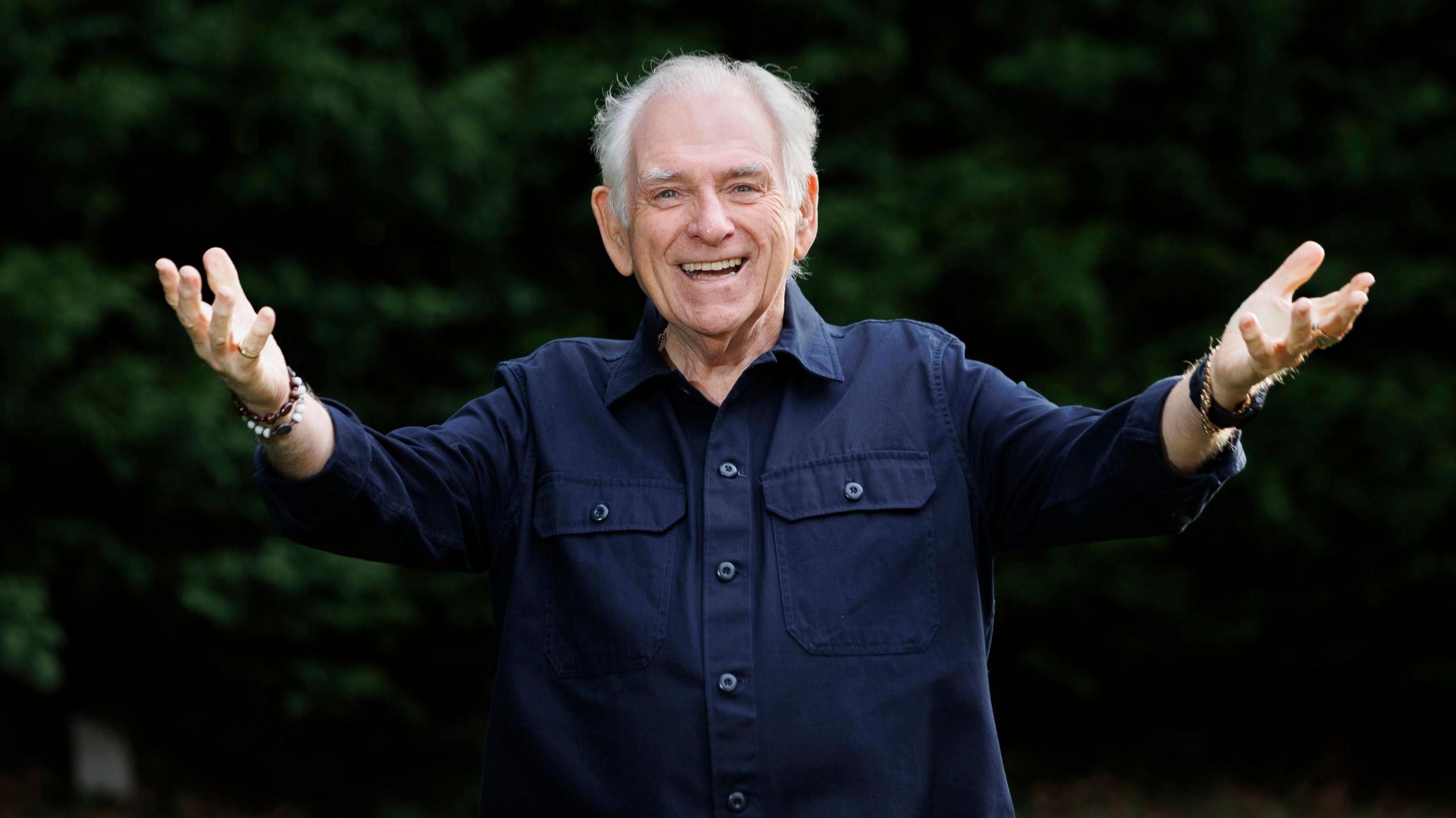 Hugo Duncan smiling at the camera with his arms outstretched. He is wearing a navy shirt. Behind him there are green bushes.