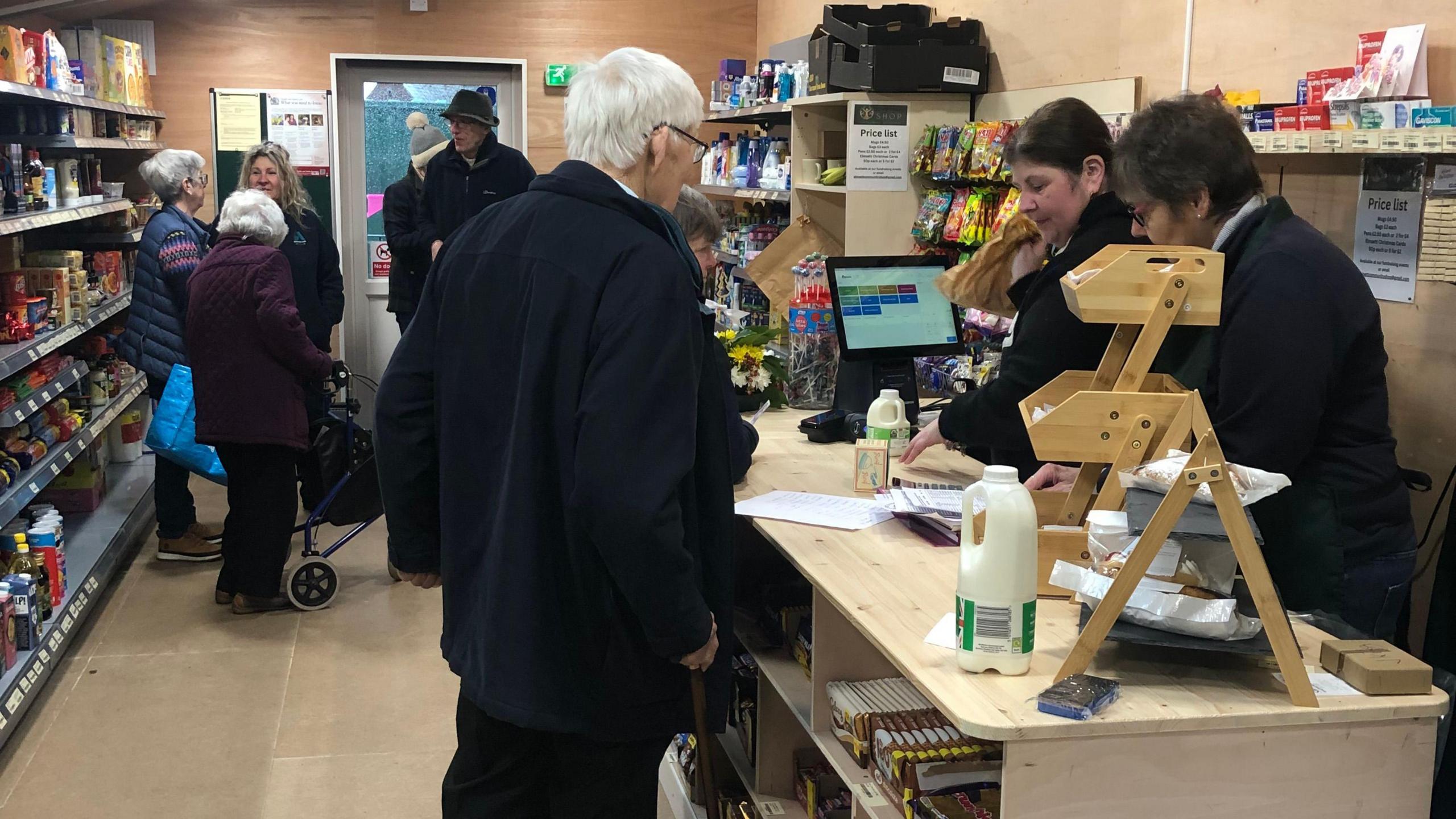 A shop scene with people standing in the background, food and tins on shelves and volunteers behind a wooden desk dealing with customers