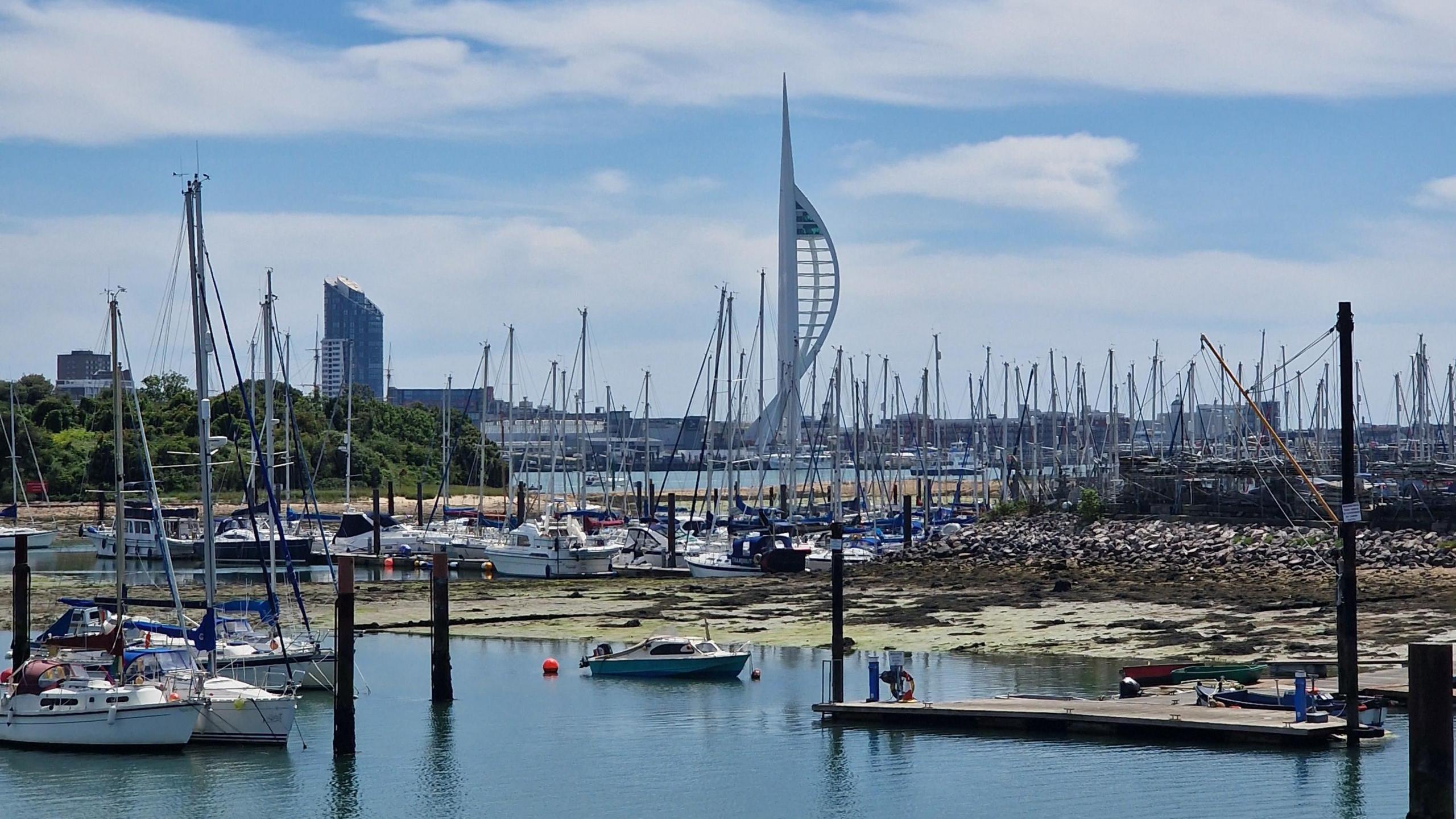 SATURDAY - The view over Gosport Harbour looking towards Portsmouth with the tall white Spinnaker Tower on the horizon. In the distance you can see the skyline of Portsmouth with a large black tower block. The foreground is full of small white yachts with their masts. The water in the foreground is blue and overhead the sky is blue with fluffy white clouds.