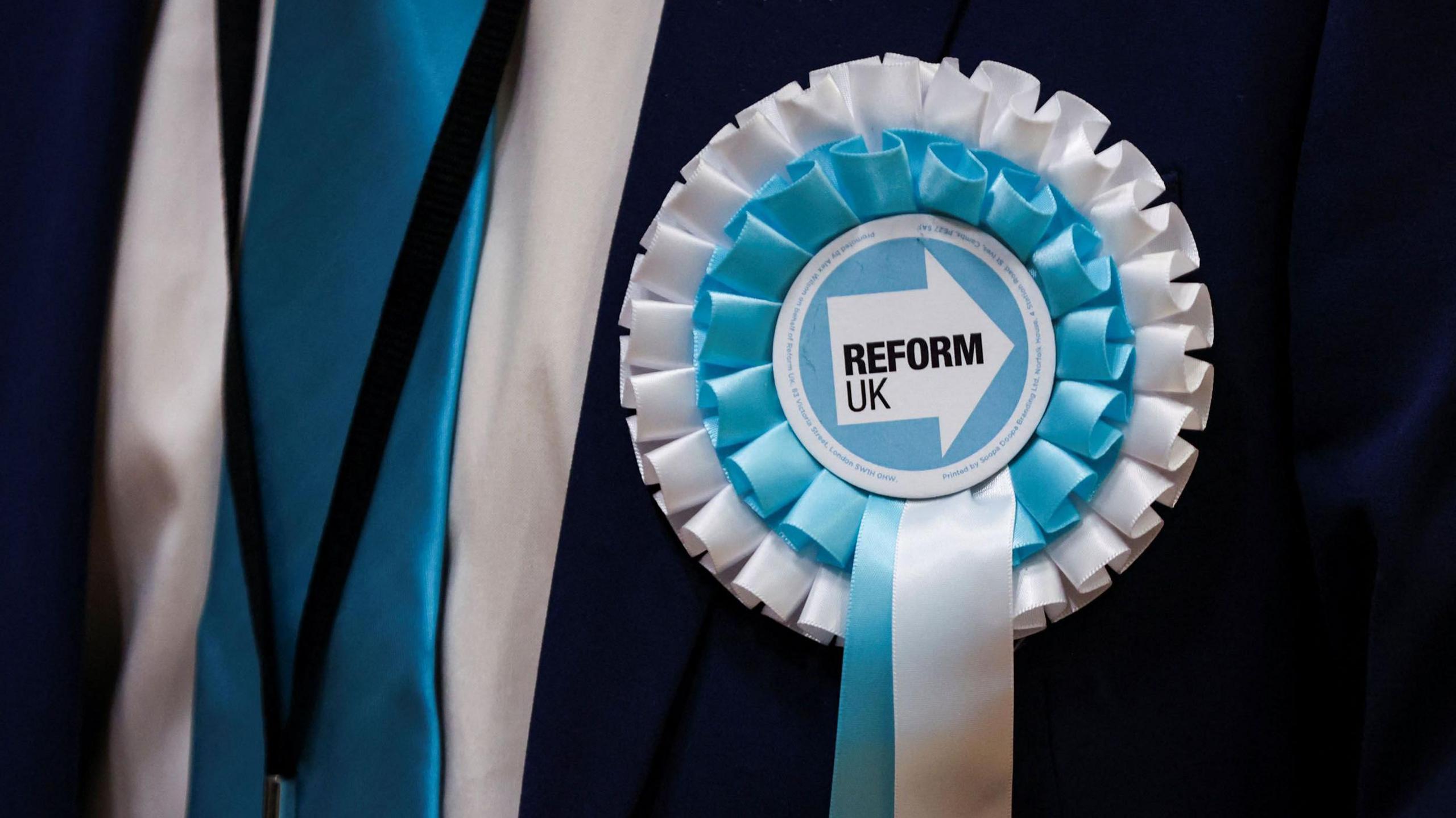 A close up picture of a Reform UK blue and white rosette attached to a suit.