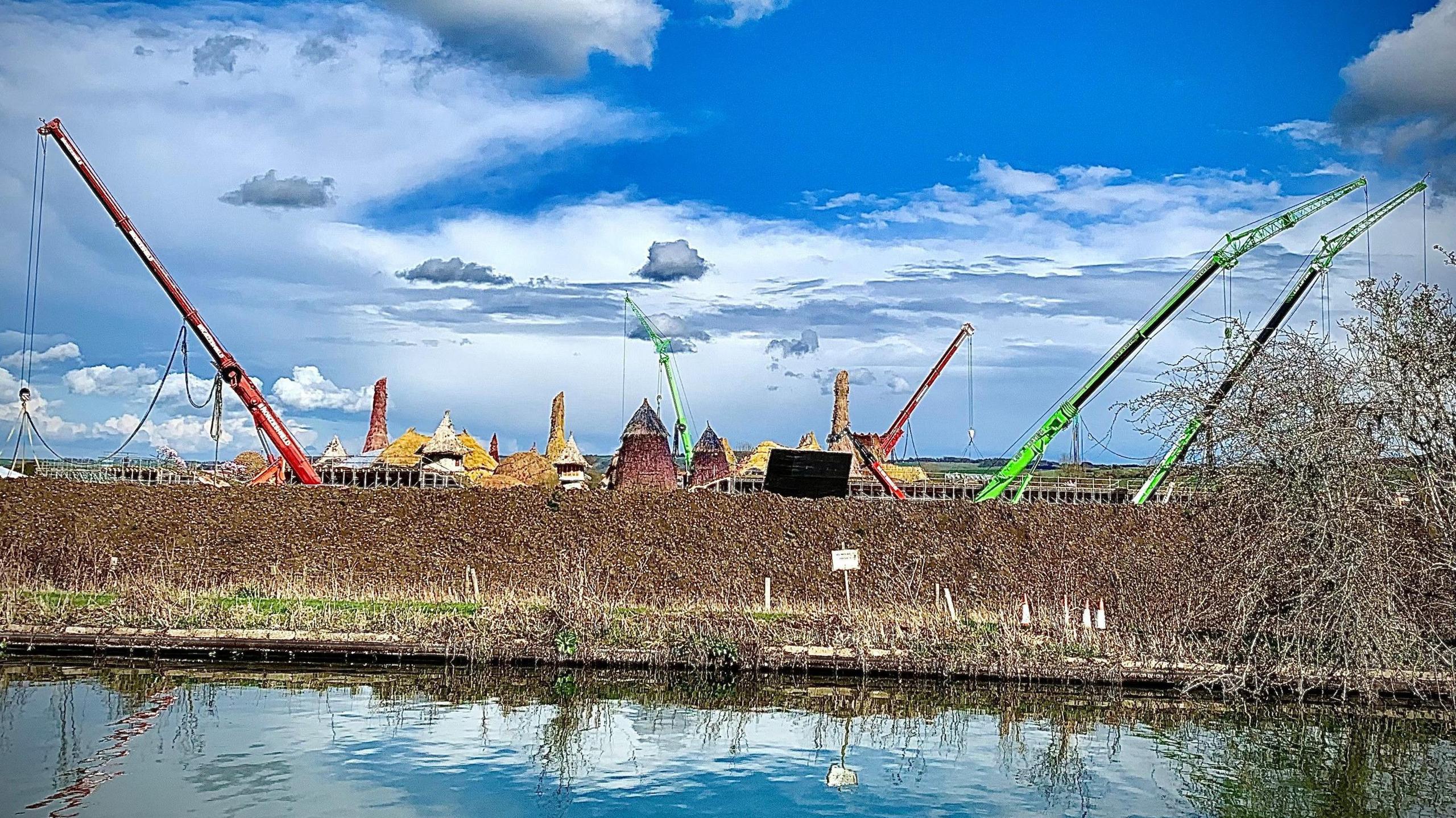 A picture taken from beside a canal and behind temporary fencing. The film set is behind the fencing, with cranes and many thatched-roof homes. They are mostly hidden by the fencing. 