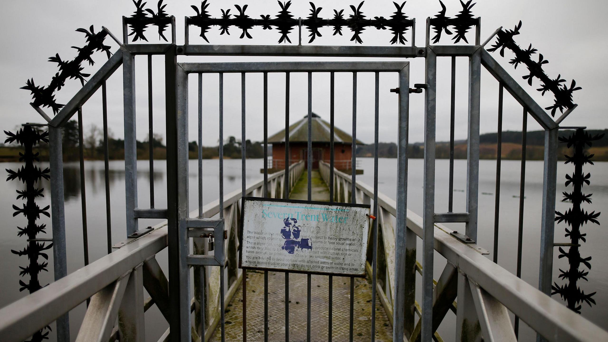 A sign hangs on a metal gate at Severn Trent Water's Cropston Reservoir. A bridge can be seen leading towards a brick building. 