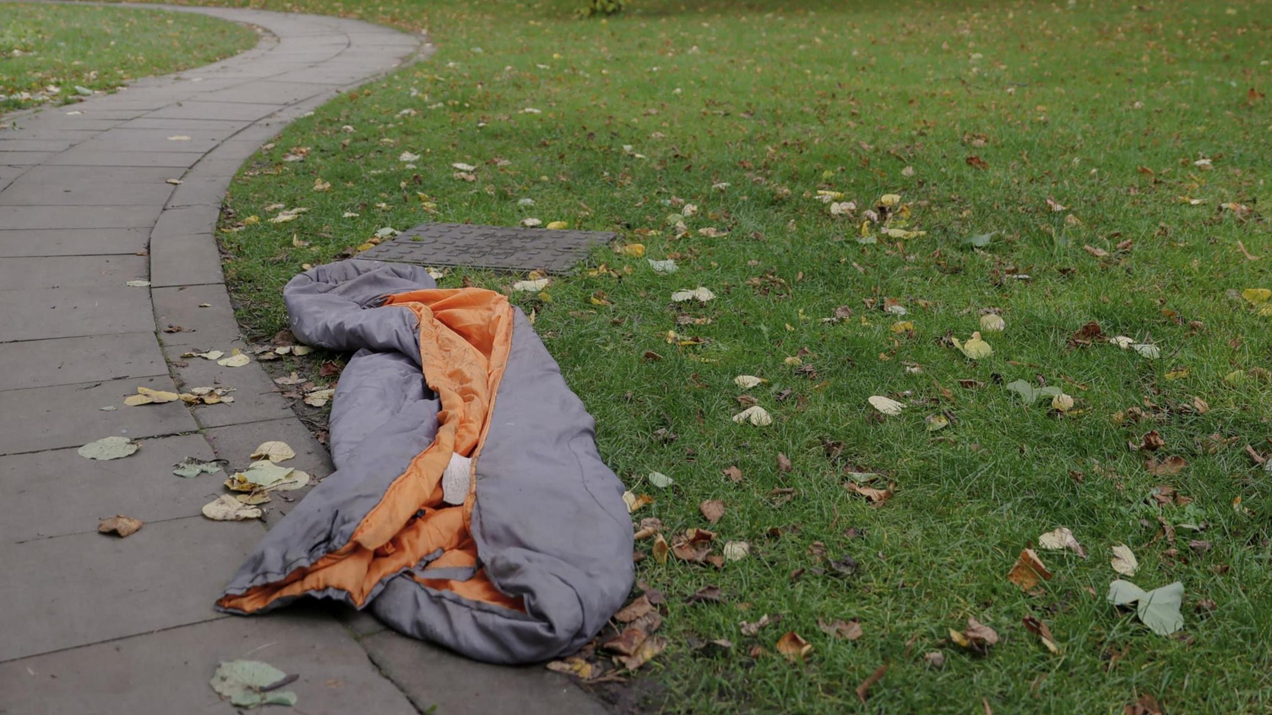 An empty grey and orange sleeping bag lies on a grassed area covered with autumn leaves. A footpath runs alongside the grass.