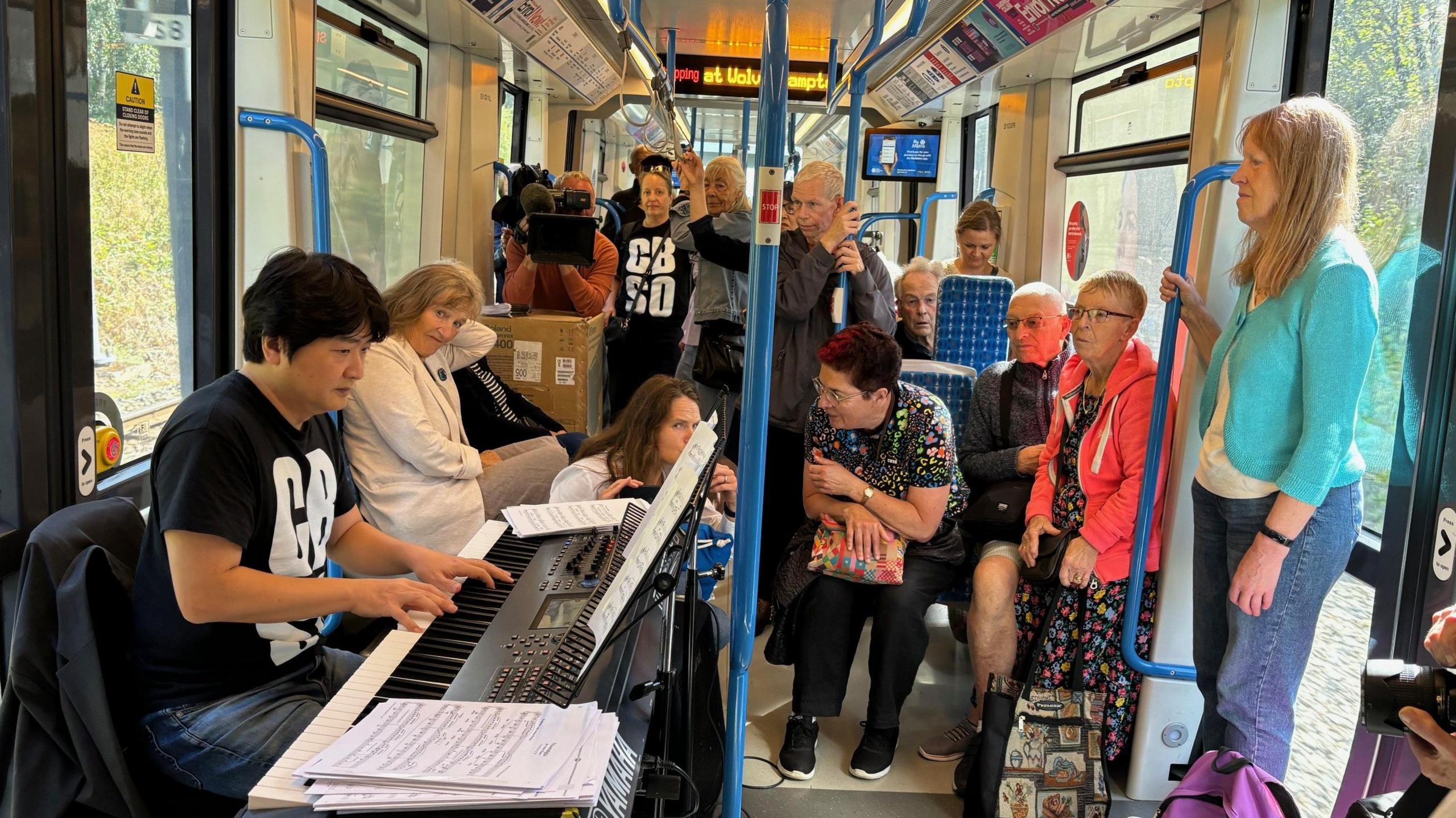 A tram full of passengers. On the left is a man performing on a keyboard, behind him someone is filming