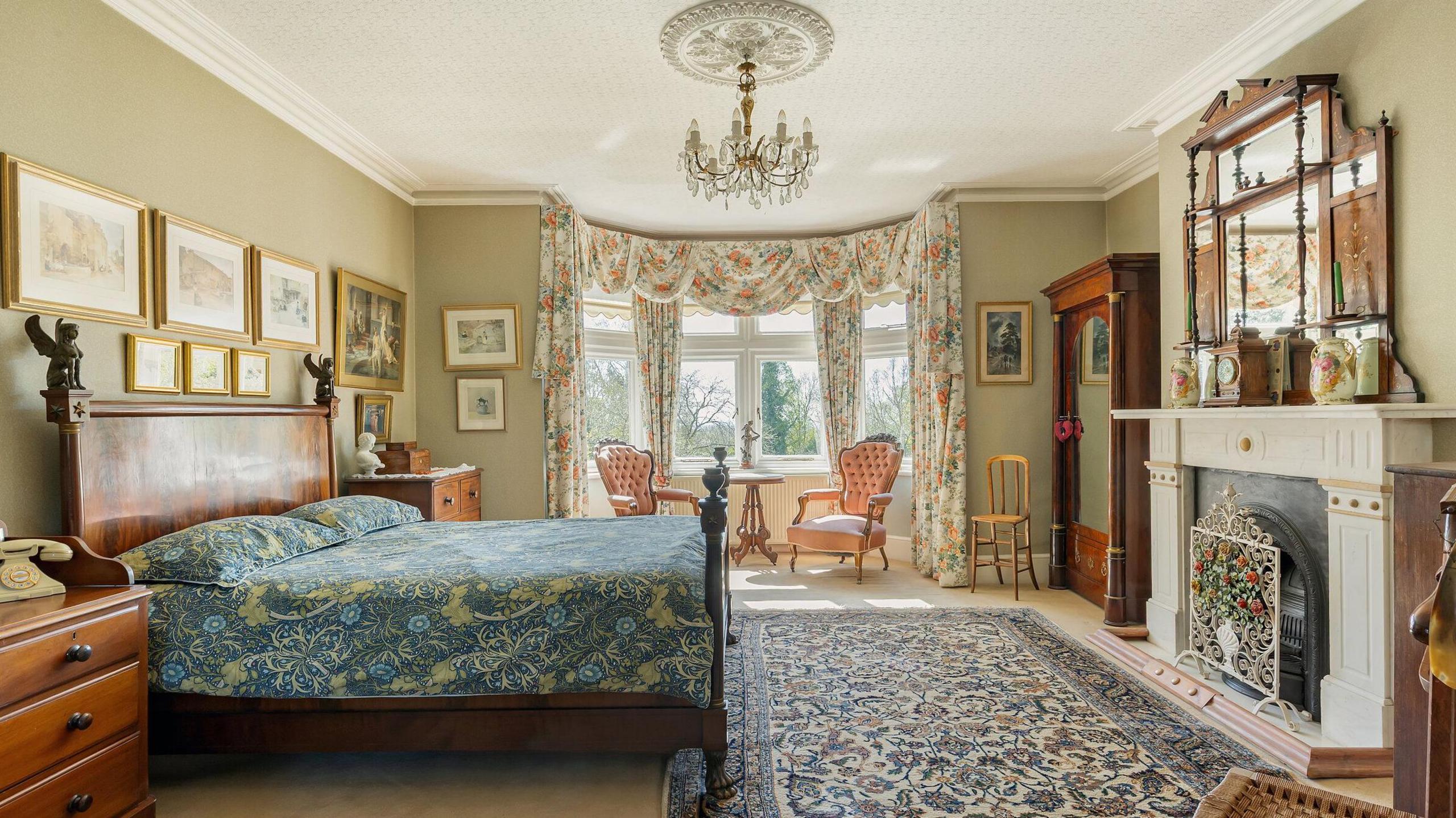 A bedroom with a fireplace and a large bay window which has two chairs in it. There are pictures on the wall and a large mirror above the fireplace