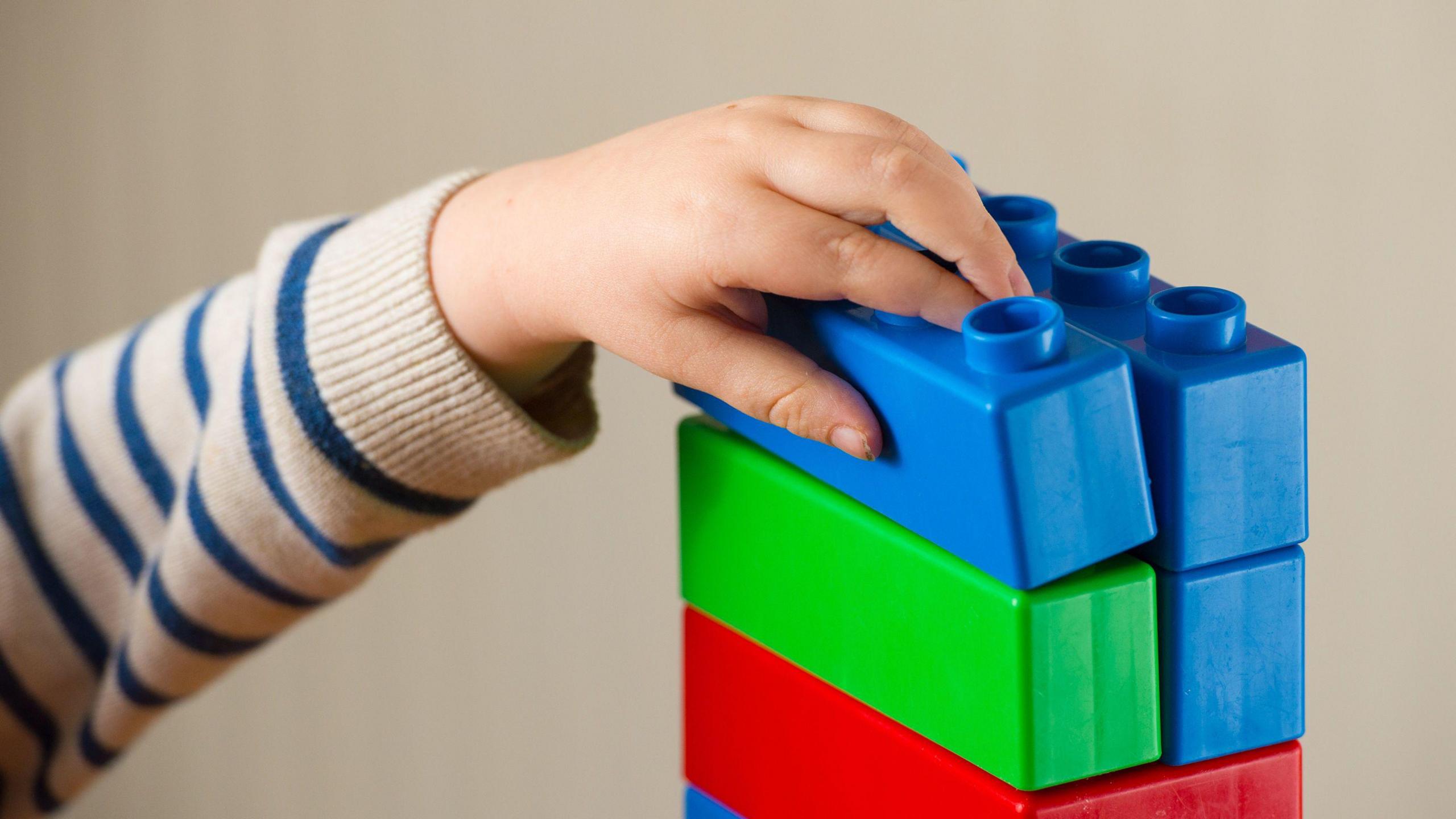 A child's arm is photographed holding a tower of large building blocks which are blue, green and red. 