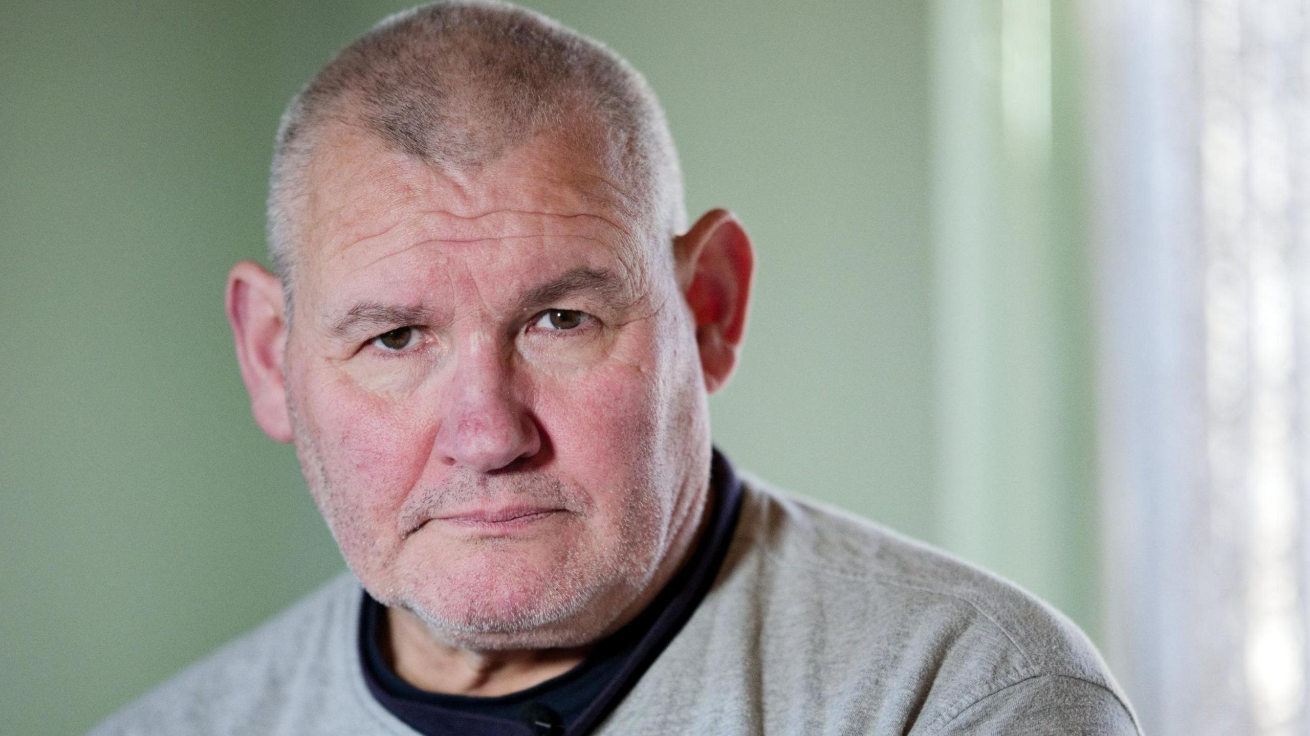 Jose looks sternly toward the camera. The wall behind him is pistachio green. He is wearing a black tee shirt and a grey sweater top with "Northfield" written on it