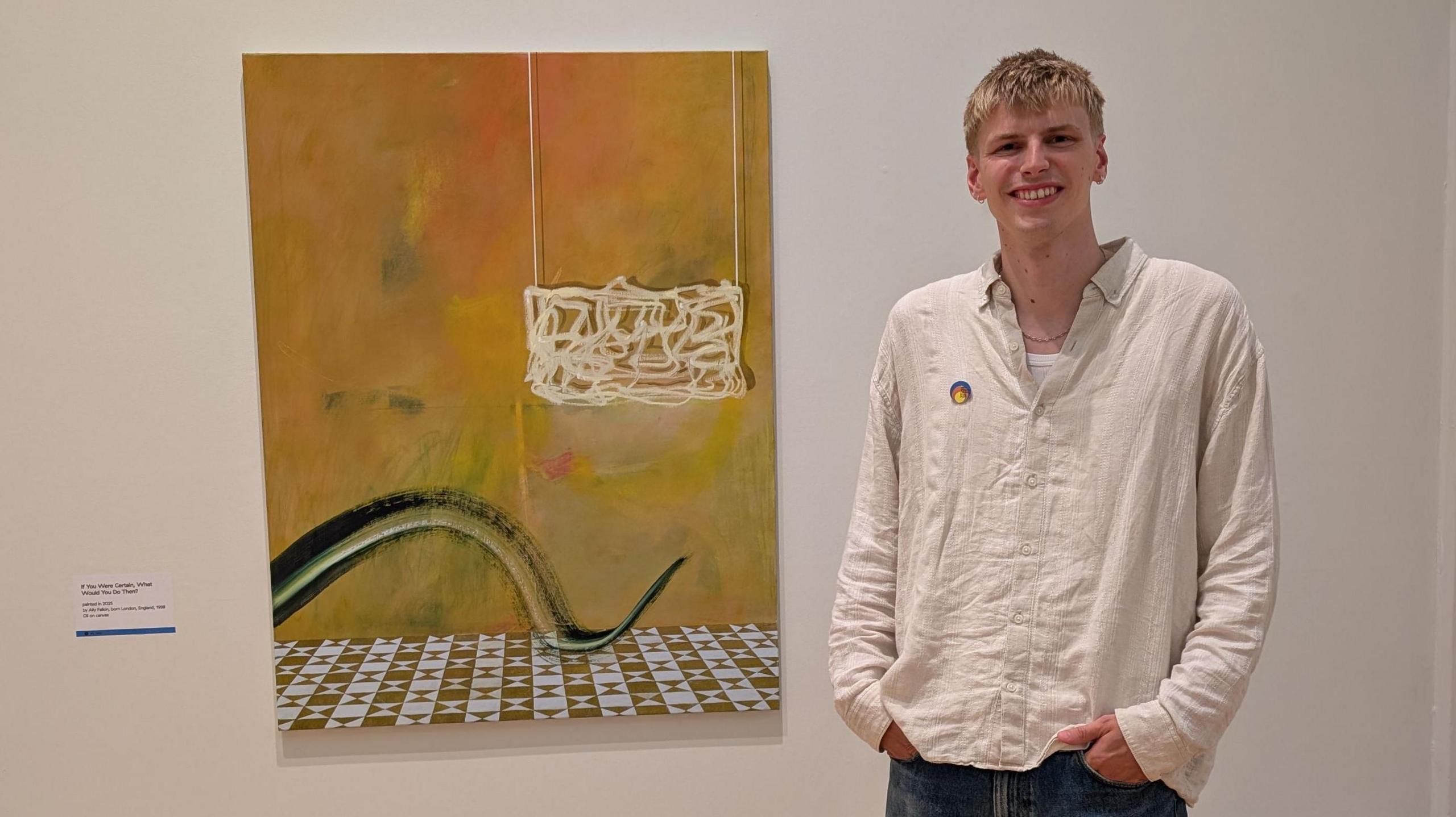 Ally Fallon has short blonde hair and is wearing a cream linen shirt and blue jeans. He is smiling at the camera as he stands next to his winning painting.