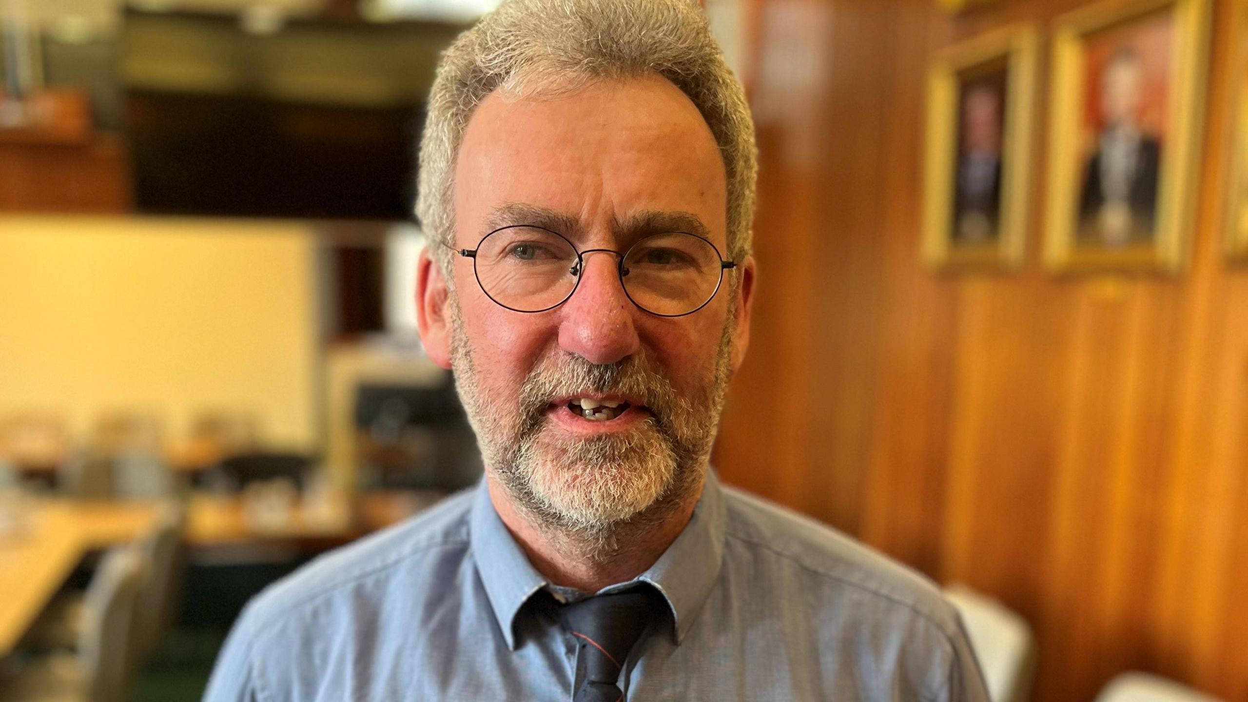 Councillor Steven Heddle, man with grey hair and beard and glasses, looking at camera, wearing a grey shirt, and a dark tie.