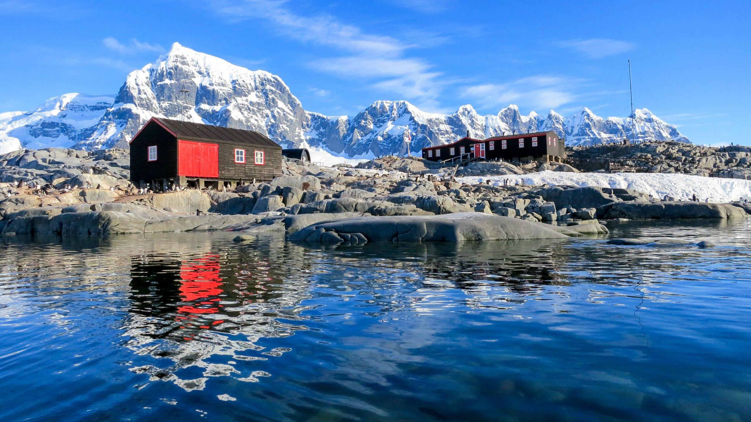 The three buildings on Port Lockroy, which are brown and red. They are in front of snow-covered mountains and reflected in the water below.