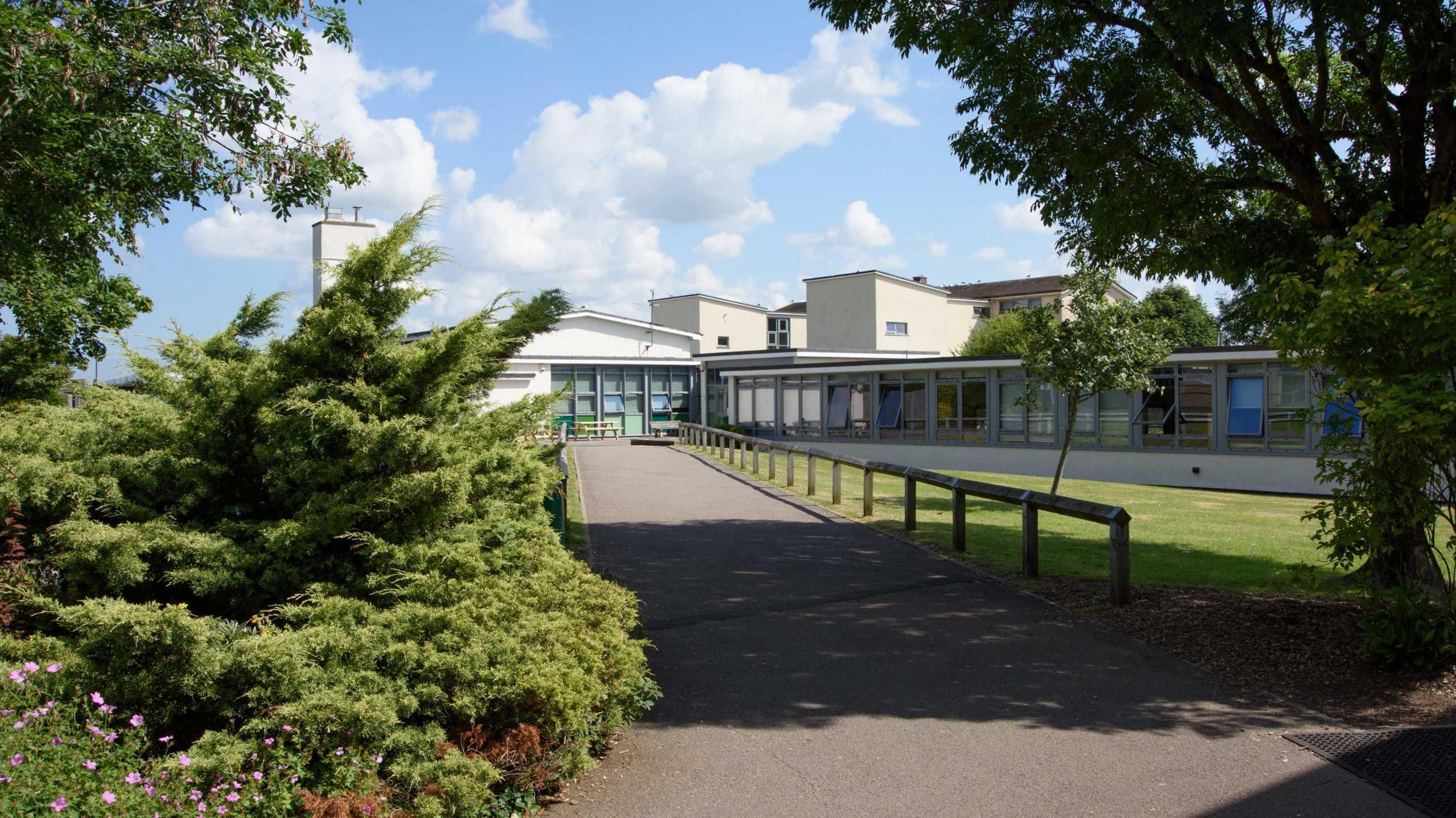 Exterior shot of Wellsway School in Keynsham. The pathway leading to an entrance to the school. There are trees either side and a lawn to the right hand side of the path.