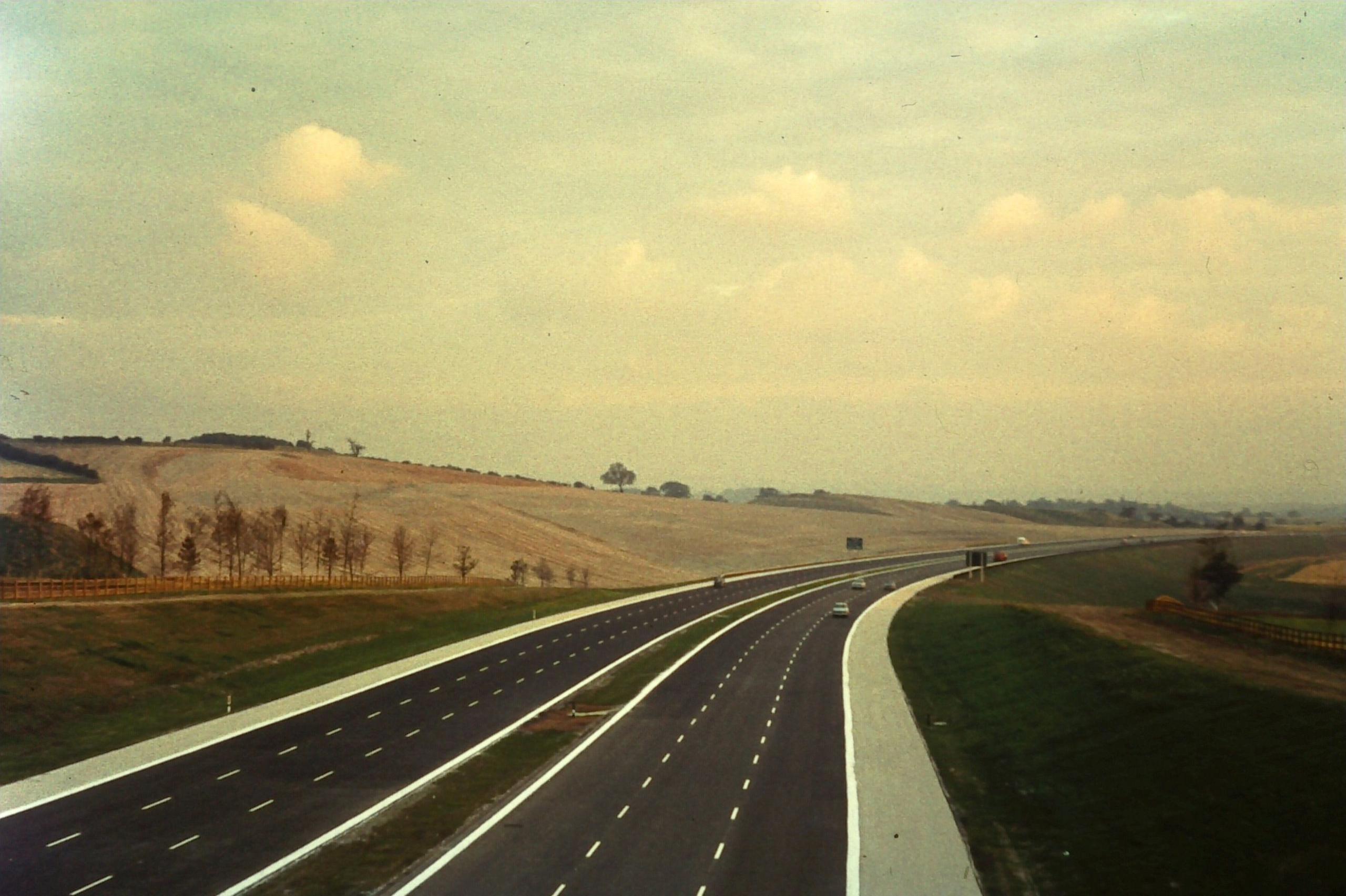 A vintage photo with a brown tinge showing a relatively empty, new motorway through countryside from the perspective of a bridge over the carriageways