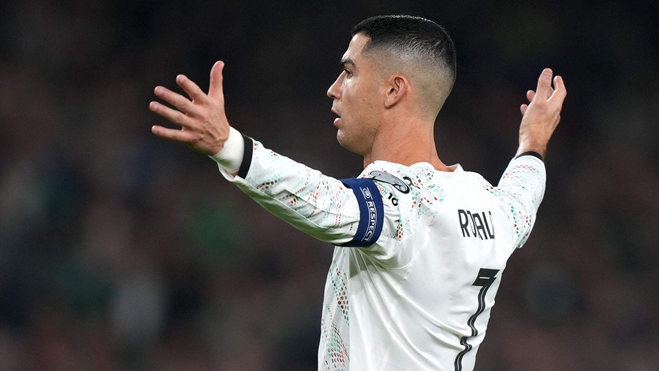 Cristiano Ronaldo with a short haircut, in a white football shit with the number seven on the back. His arms are outstretched and he has a dark blue captain's armband on his left arm.