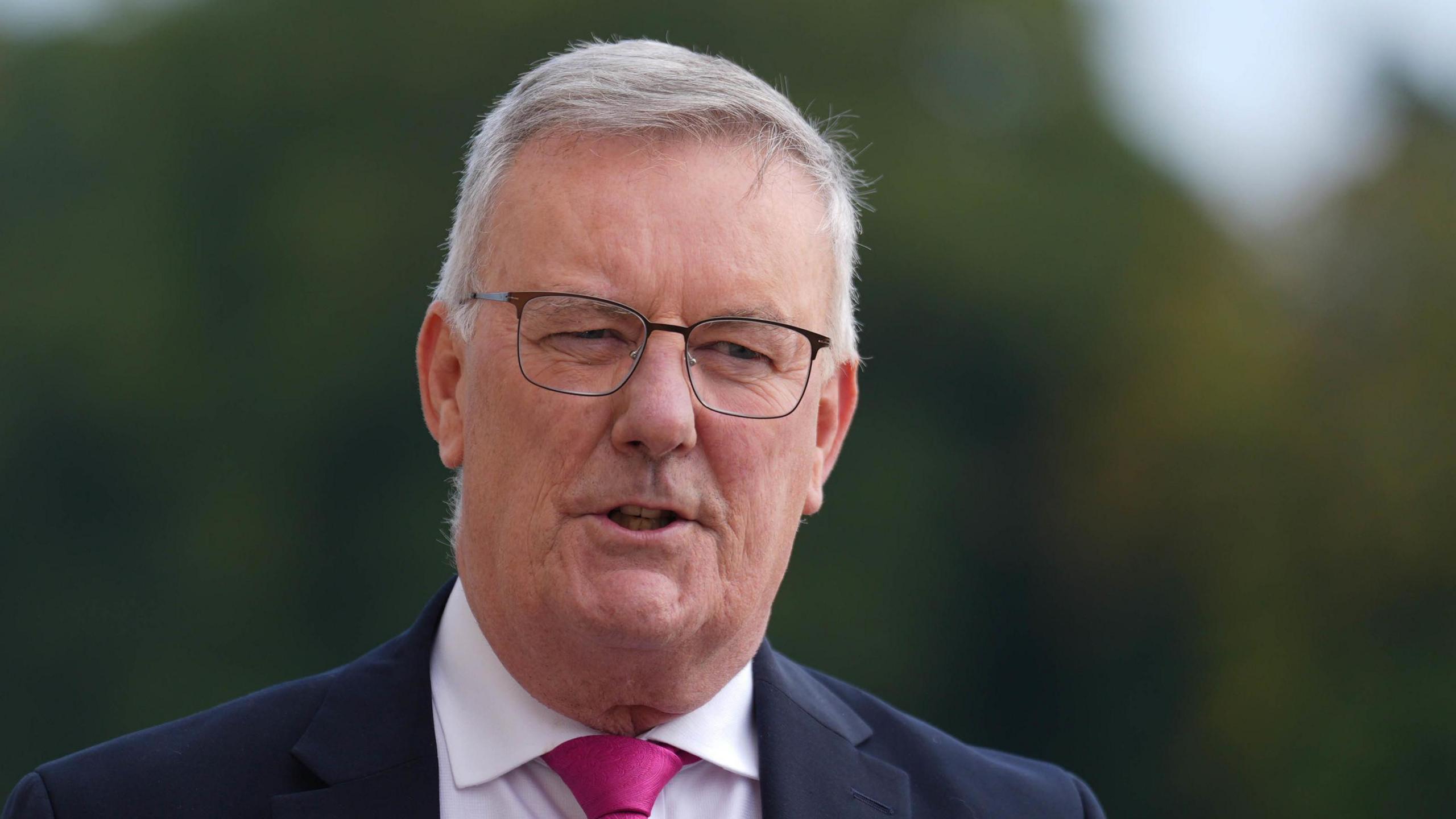 A man with short grey to white hair directs his gaze off camera, while poised to speak. He is wearing a dark blue suit jacket, white shirt, purple tie and glasses.